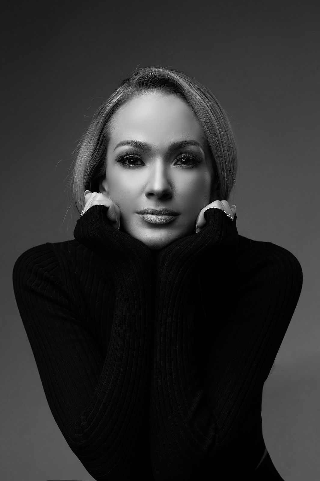Elegant blonde woman in black turtleneck posing with hands framing face on gray background.