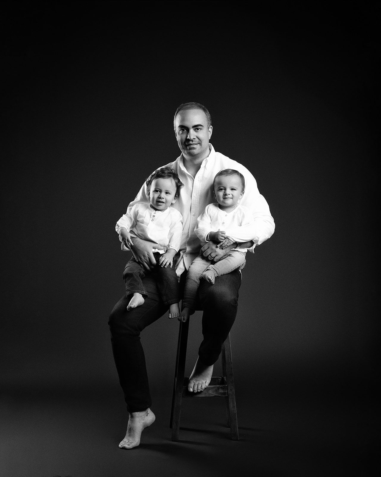 Black and white studio portrait of father sitting on stool holding two babies in white outfits.
