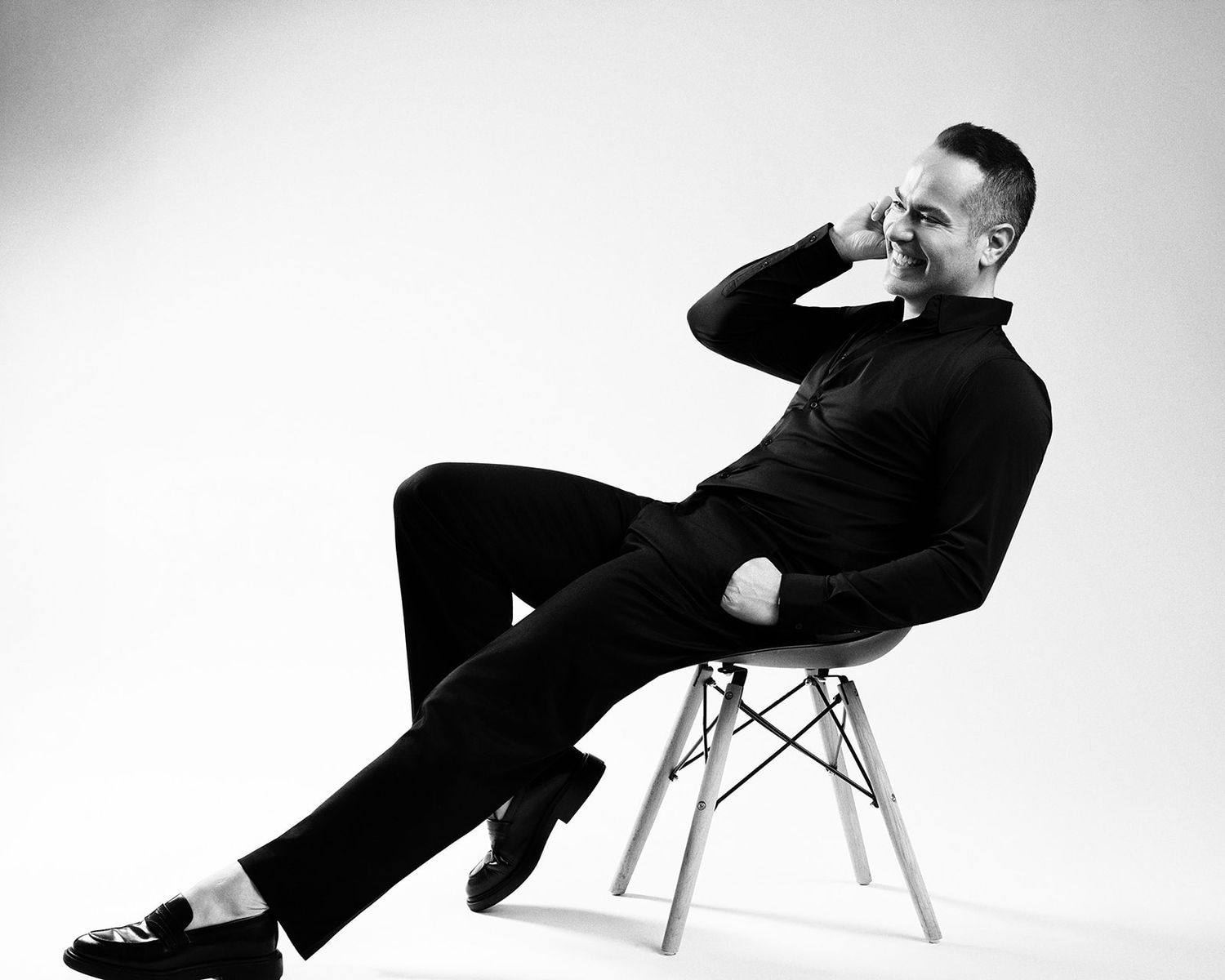 Black and white photo of a man in all-black outfit sitting relaxed on a modern chair, hand raised to face.