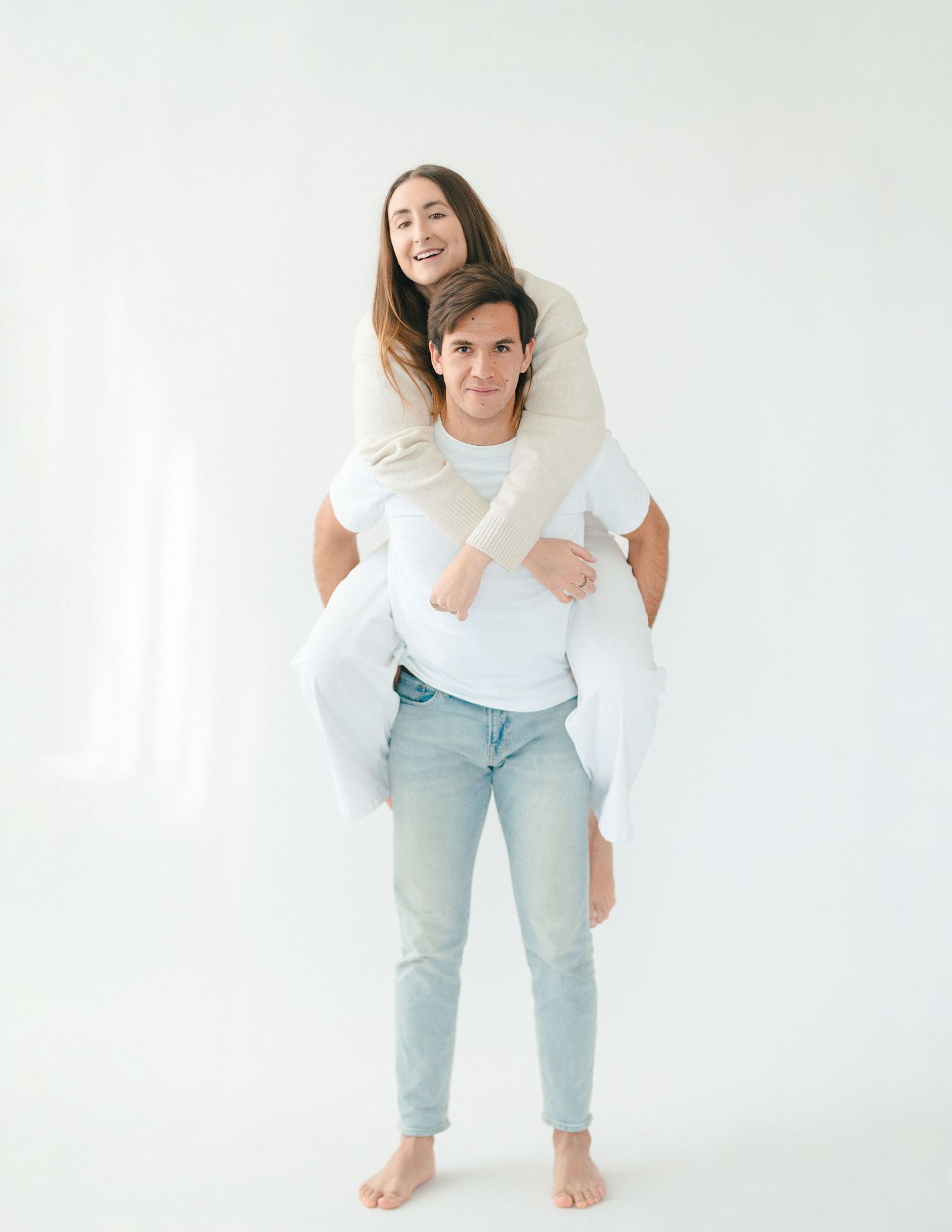 A smiling couple posing playfully with the man giving the woman a piggyback ride in a white studio.