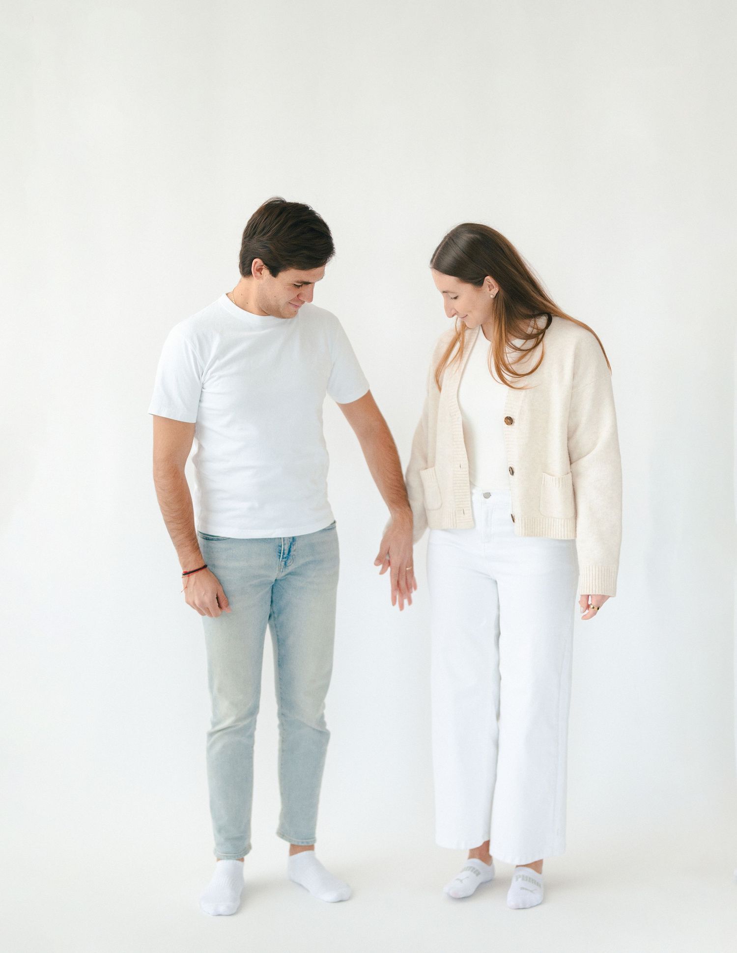 Couple in casual white outfits sharing a tender moment looking down at their hands in a white studio.