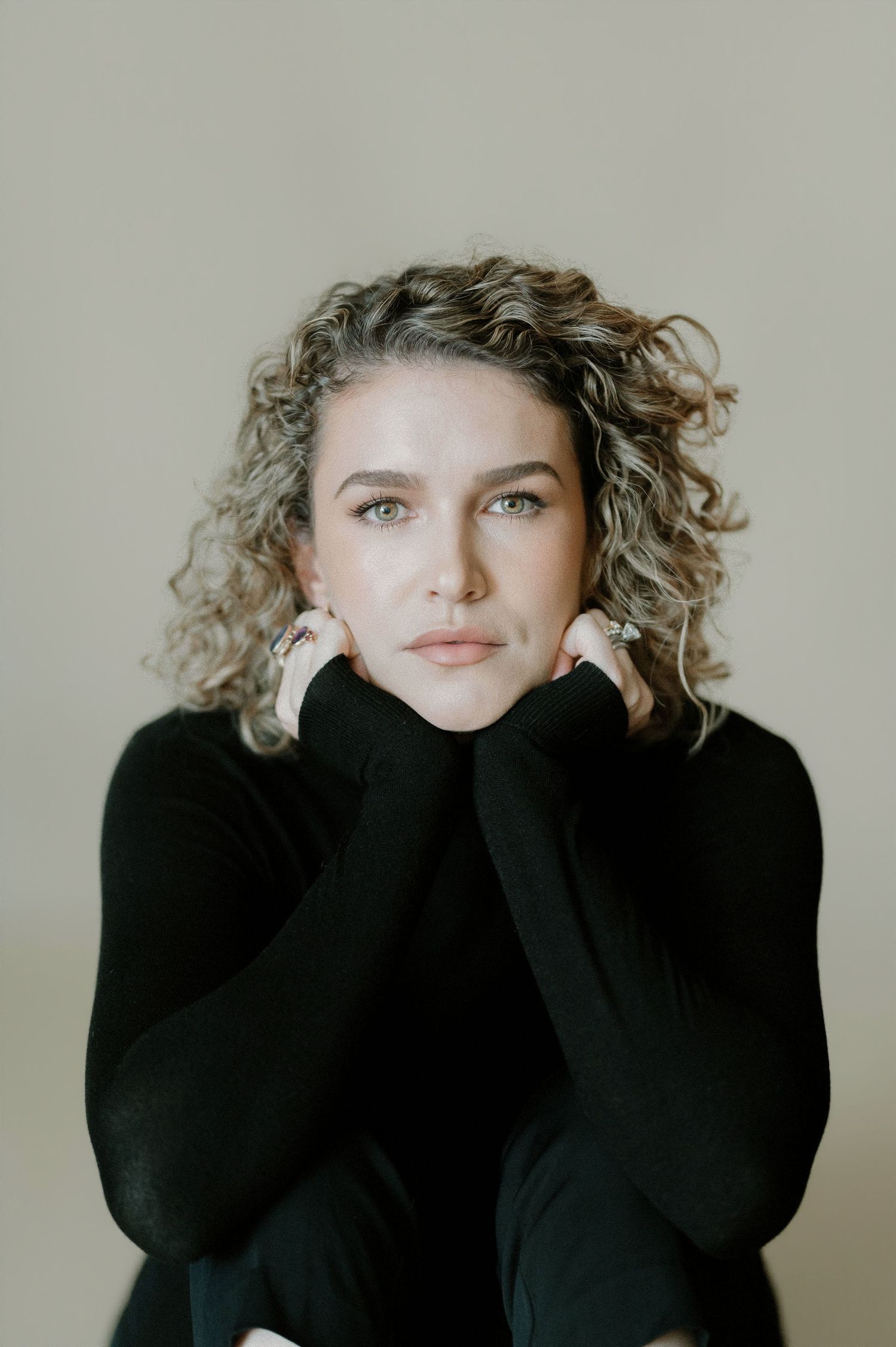 Curly-haired woman in black turtleneck rests chin on hands, close-up portrait against neutral background.