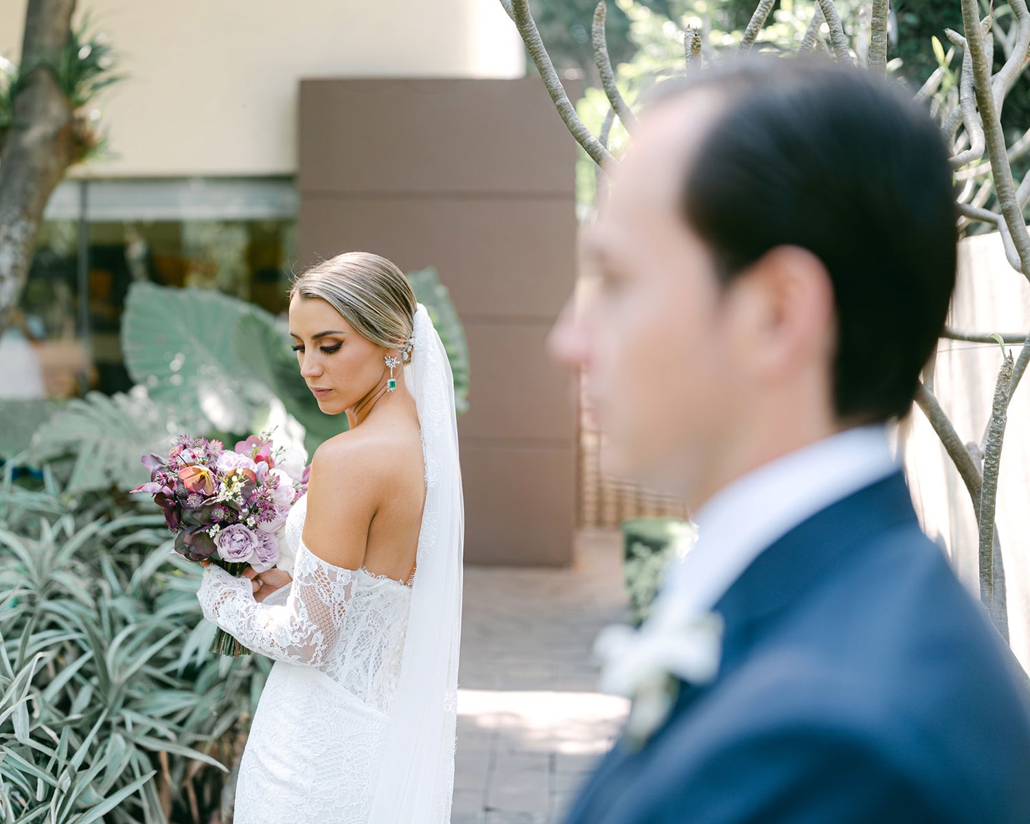 Bride in off-shoulder lace gown holding a floral bouquet with groom in foreground outdoors.