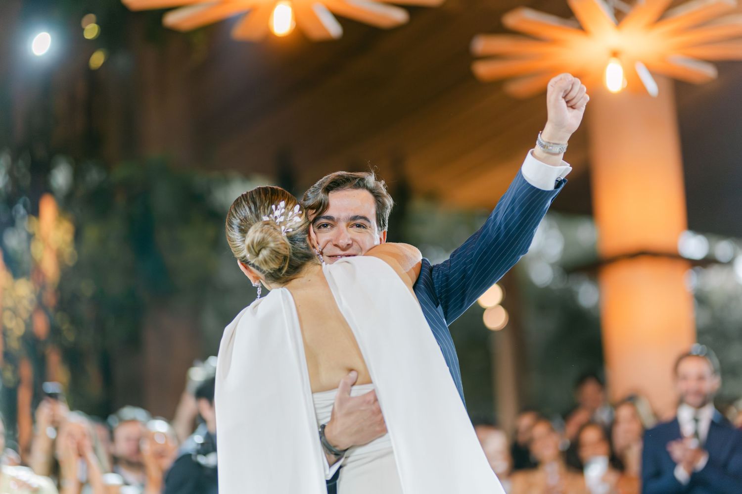 Groom in blue suit lifts bride in white gown while celebrating joyfully at wedding reception.