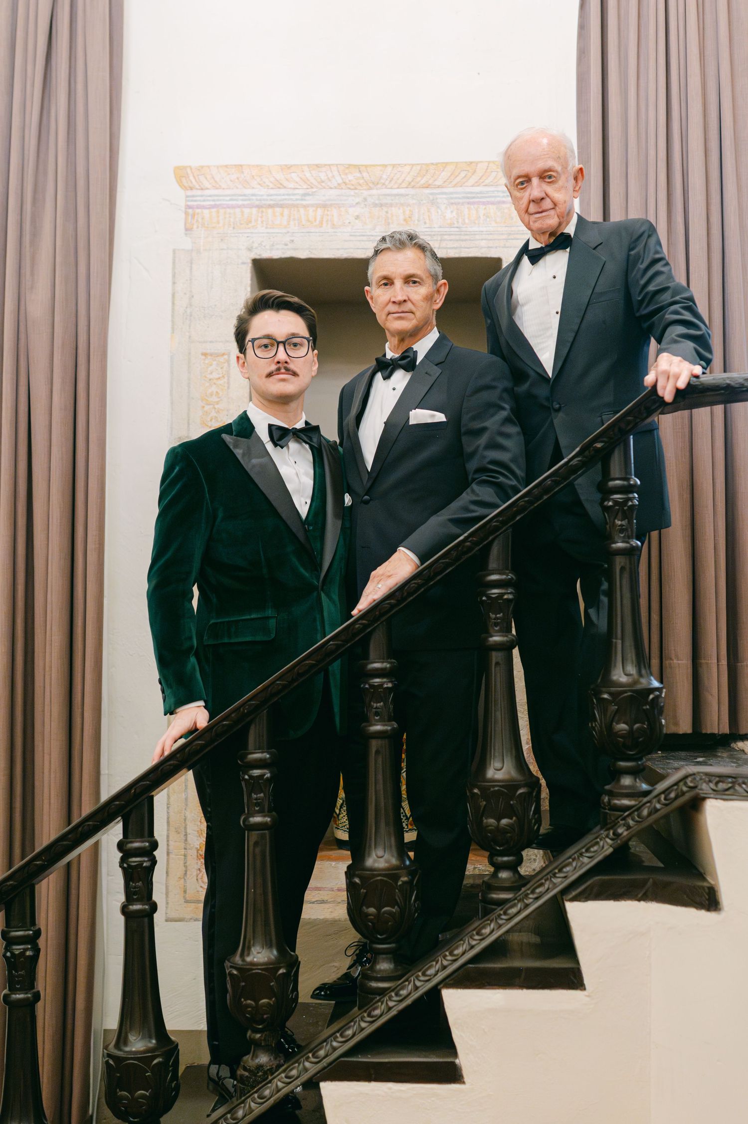 Three men in tuxedos posing elegantly on a grand staircase at a formal wedding venue.