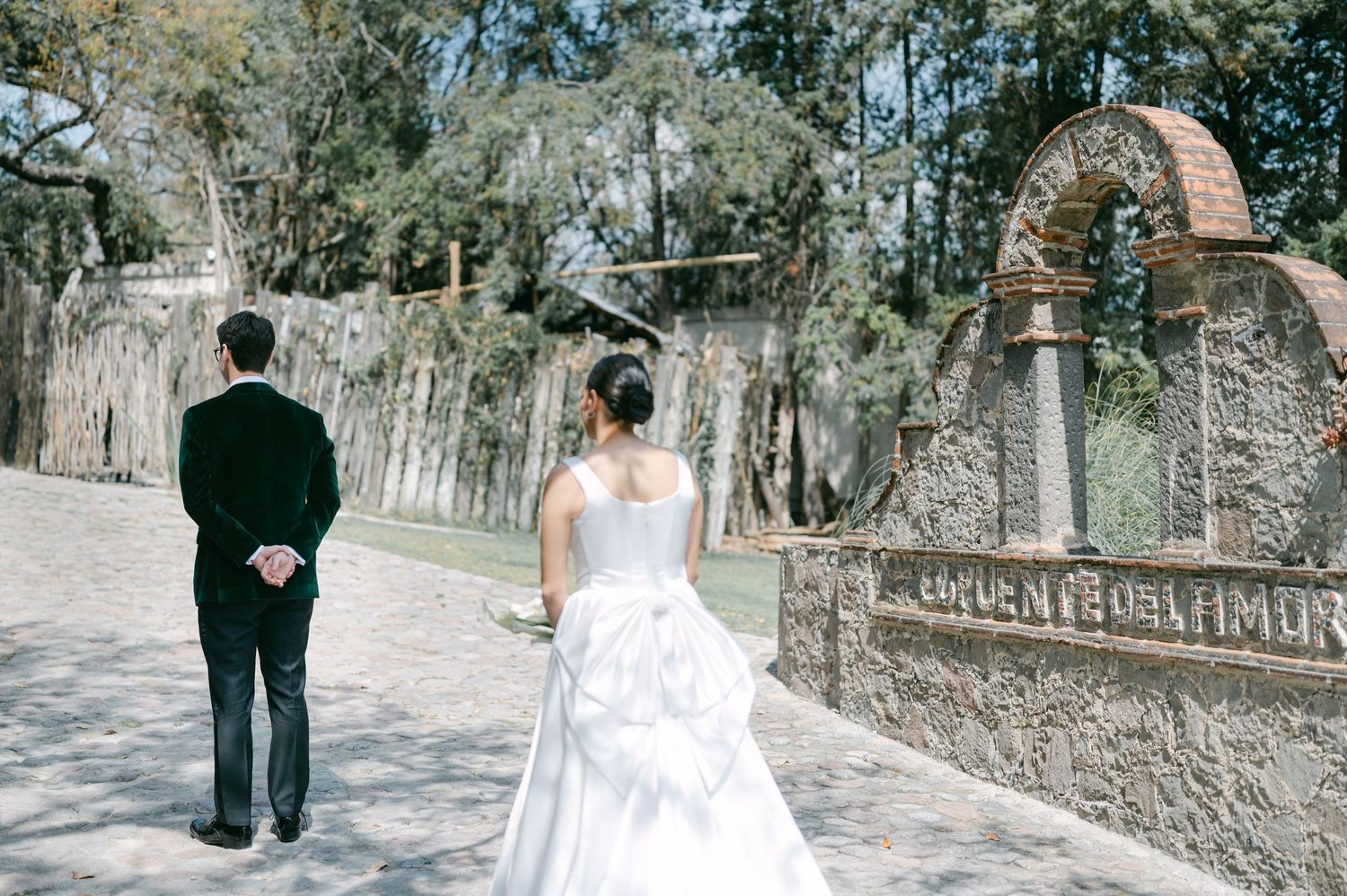 Bride and groom walk away hand in hand toward ornate stone arch monument along a sunlit garden pathway.