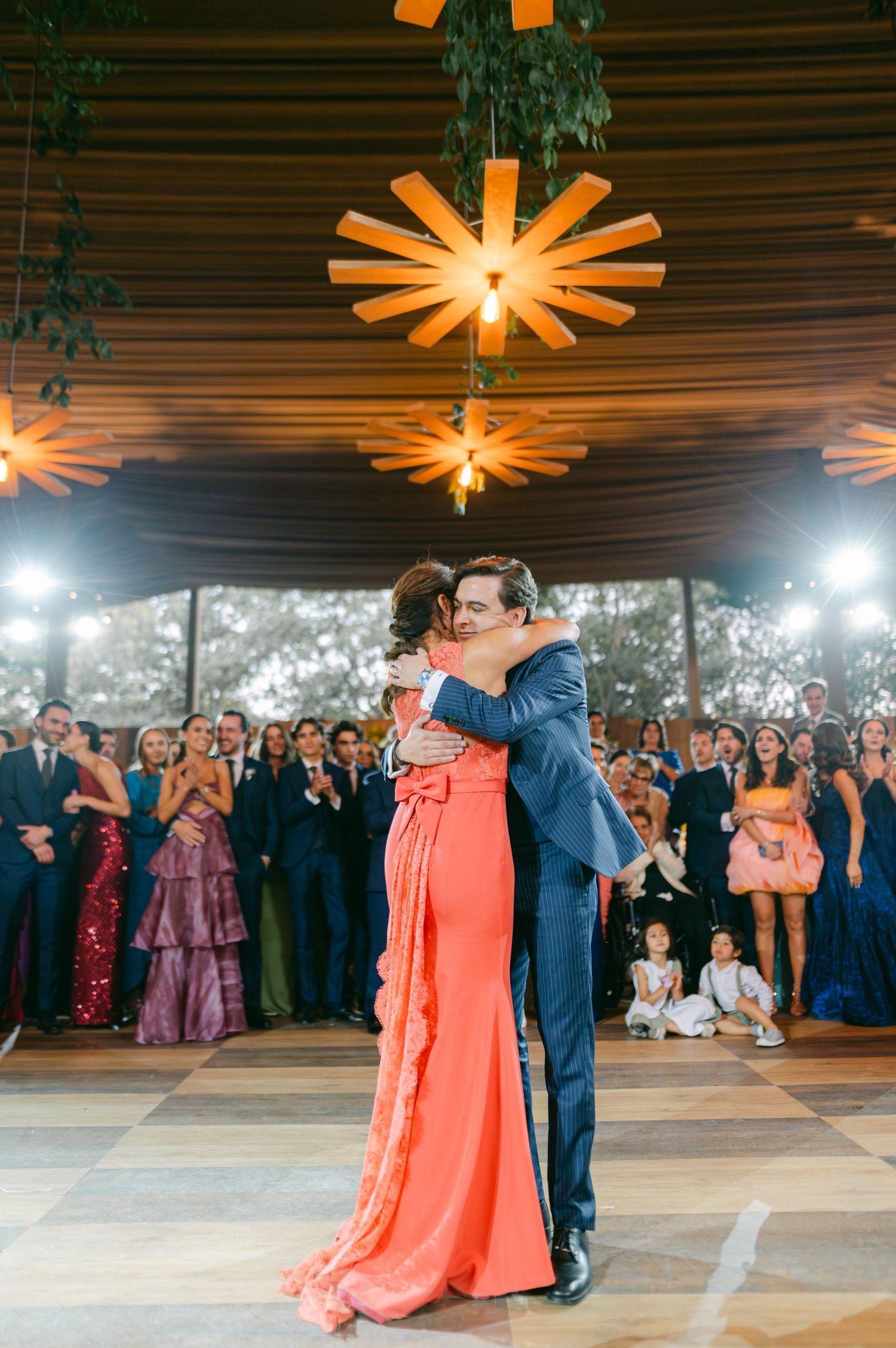 Bride in coral gown shares first dance with groom in blue suit under wooden star chandeliers at elegant wedding reception.
