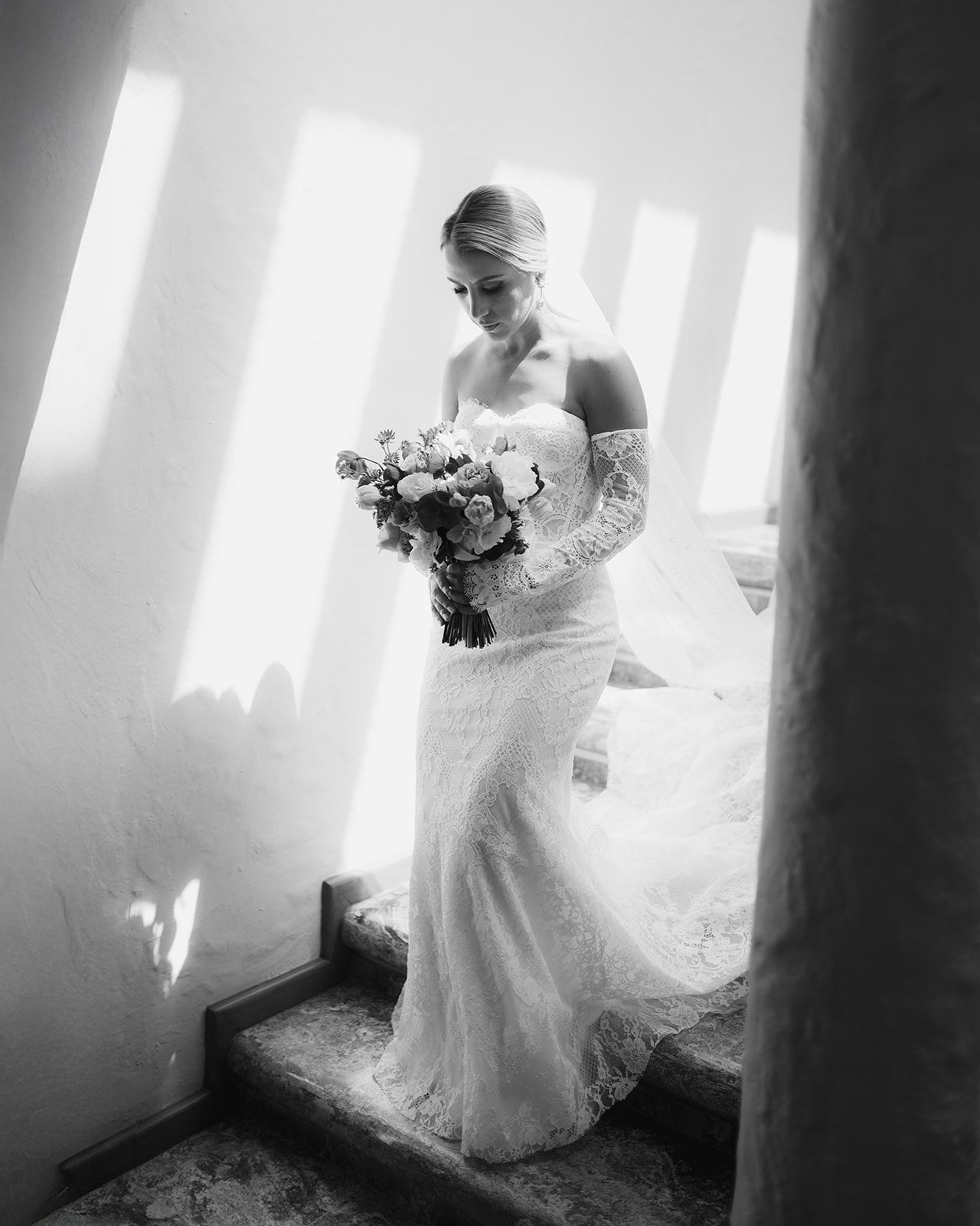 Elegant bride in lace off-shoulder gown holding floral bouquet on staircase in black and white.