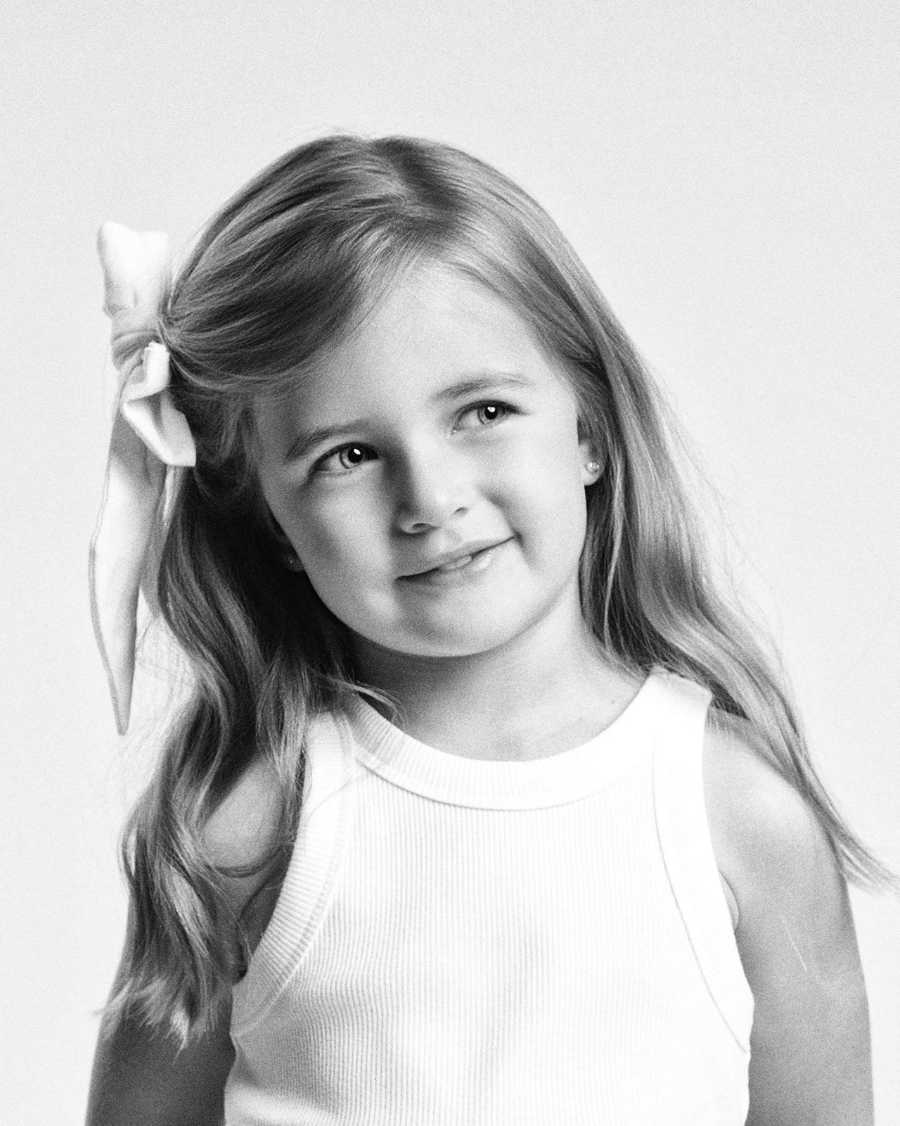 Close-up black and white portrait of a smiling young girl with long hair and a white bow accessory.