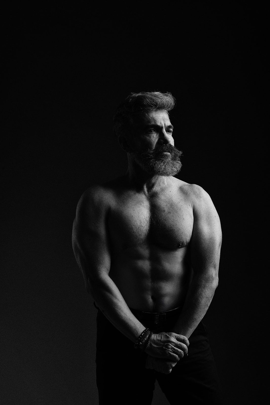 Black and white photo of a muscular shirtless bearded man posing against a dark background.