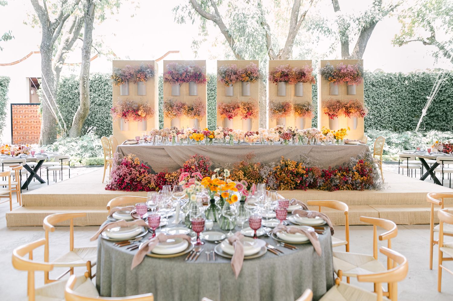 Elegant outdoor wedding reception with colorful floral wall backdrop and round tables with gray linens.