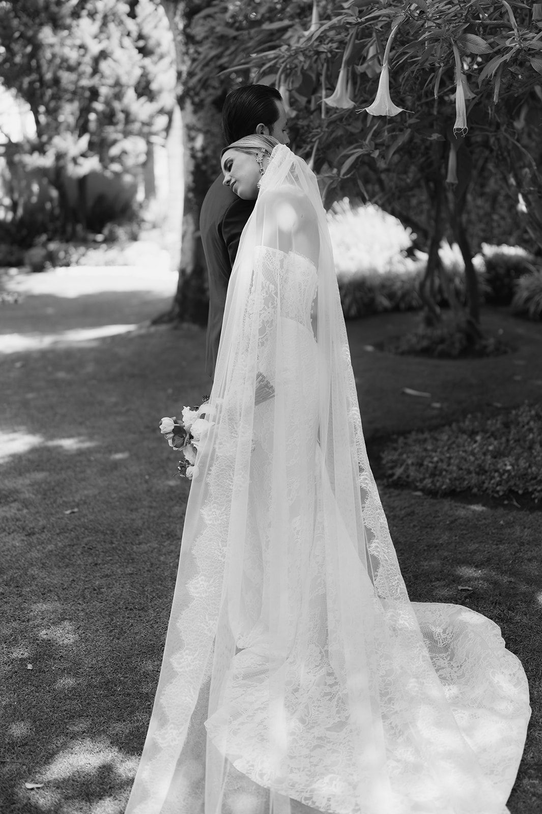 Black and white photo of bride and groom embracing in a garden, bride wearing a lace gown with cathedral veil.