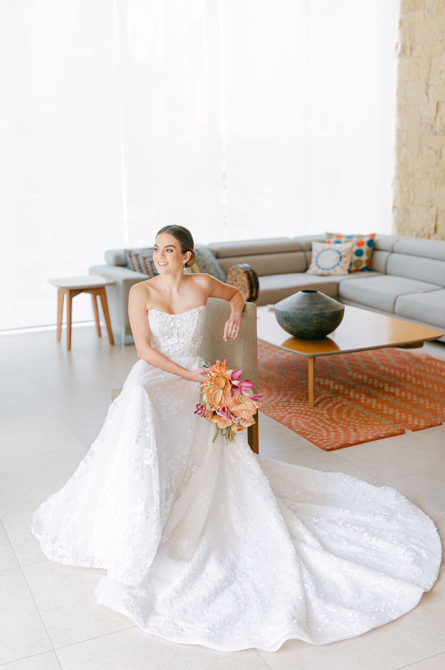 Elegant bride in strapless lace ball gown seated holding colorful tropical bridal bouquet indoors.