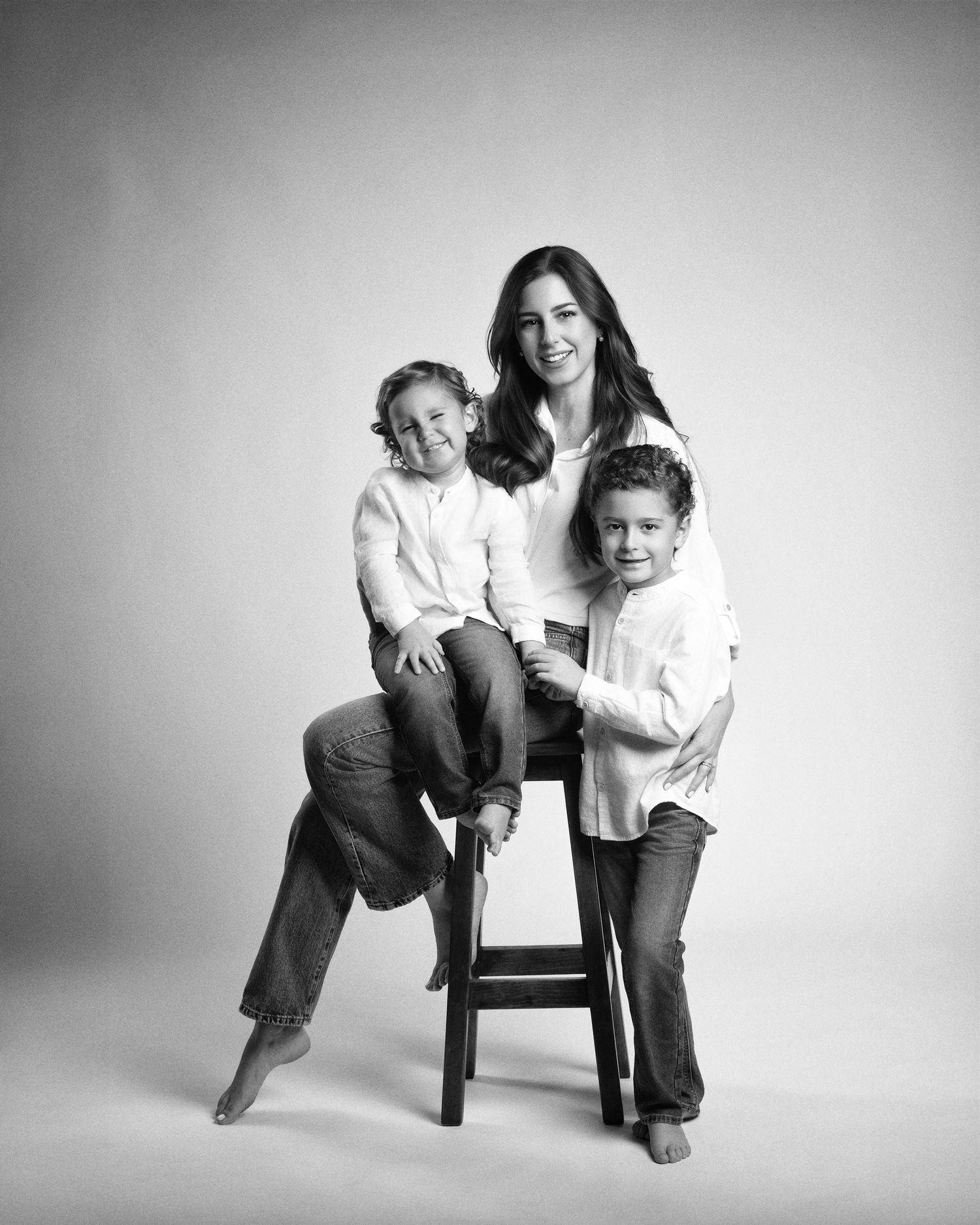 Black and white studio portrait of a mother sitting on a stool with two young children posing together.