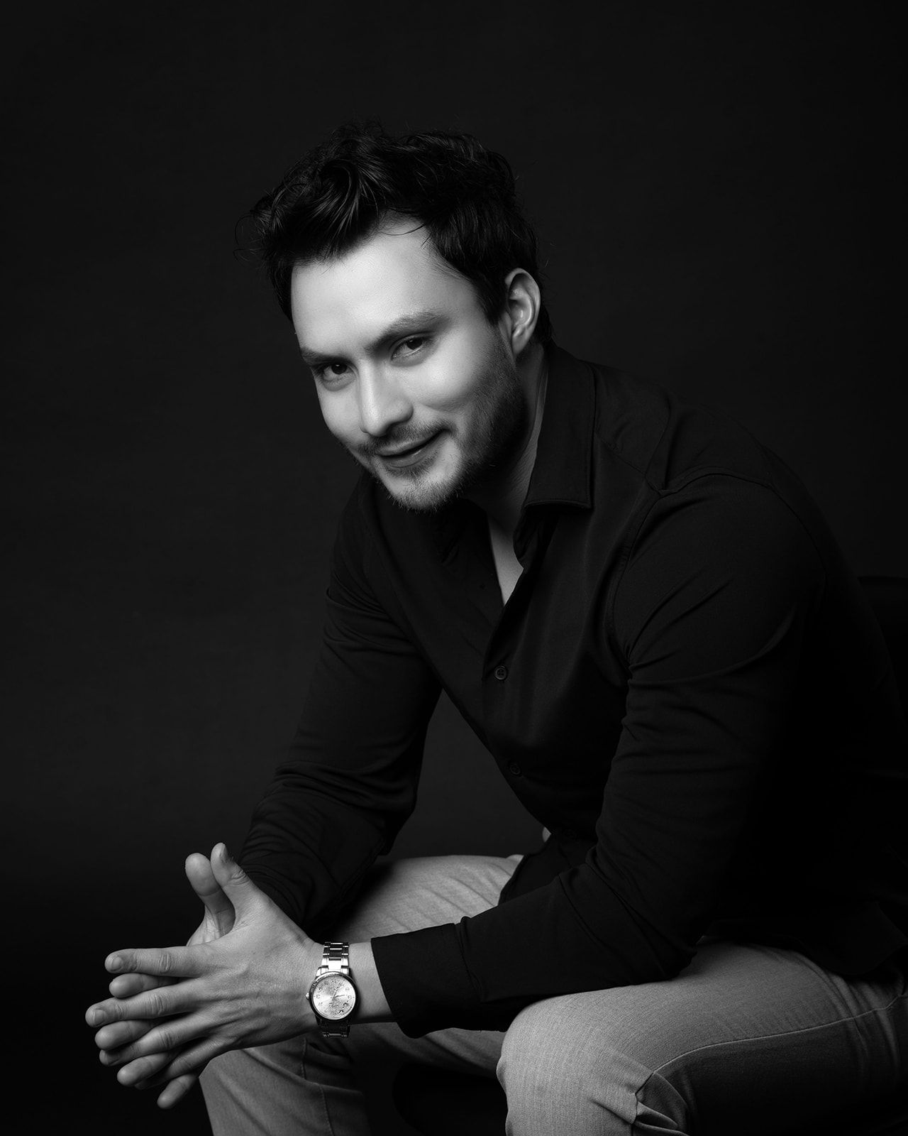 Black and white portrait of a smiling man in a black shirt wearing a watch, seated casually.
