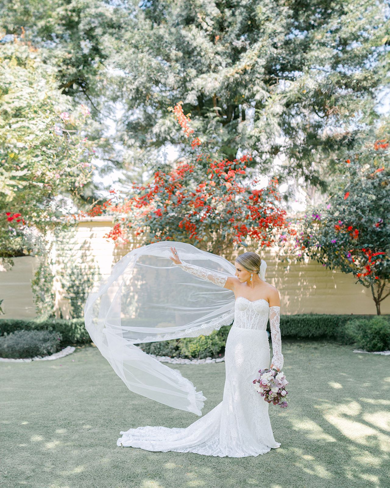 Bride in lace wedding gown twirling cathedral veil in garden with vibrant red flowering trees.