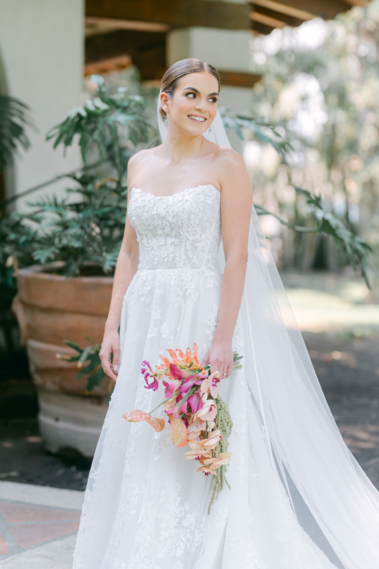 Elegant bride in strapless lace gown holding colorful tropical bouquet outdoors at wedding venue.