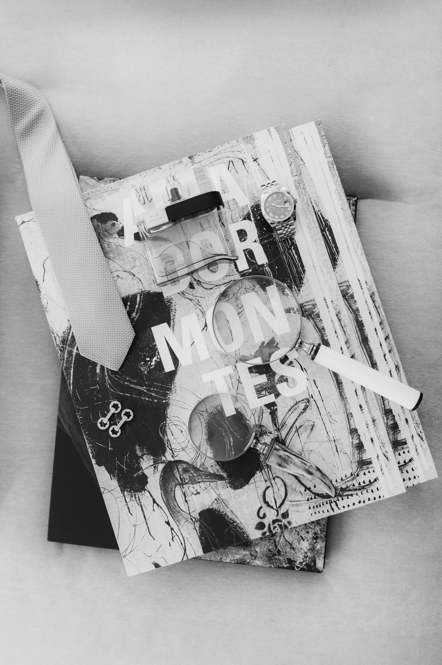 Black and white flat lay of groom accessories including cologne, watch, and magnifying glass on decorative book.