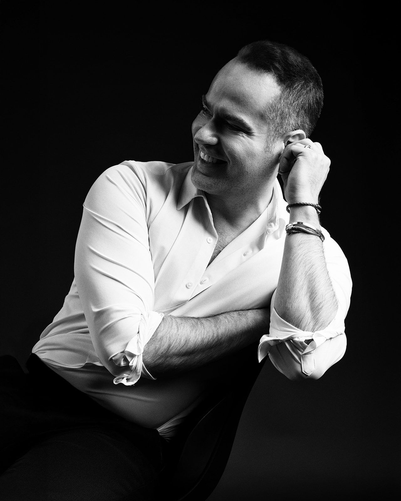 Black and white portrait of a smiling man in a white shirt, hand near face, seated against dark background.