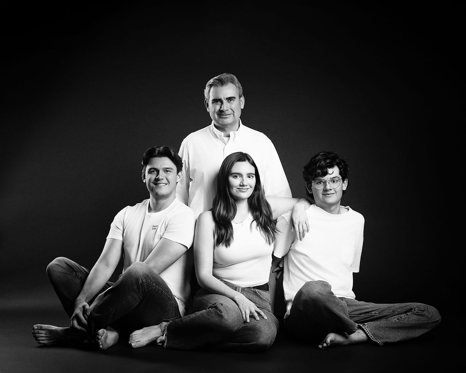 Black and white family portrait of father and three teens posing on dark studio background.