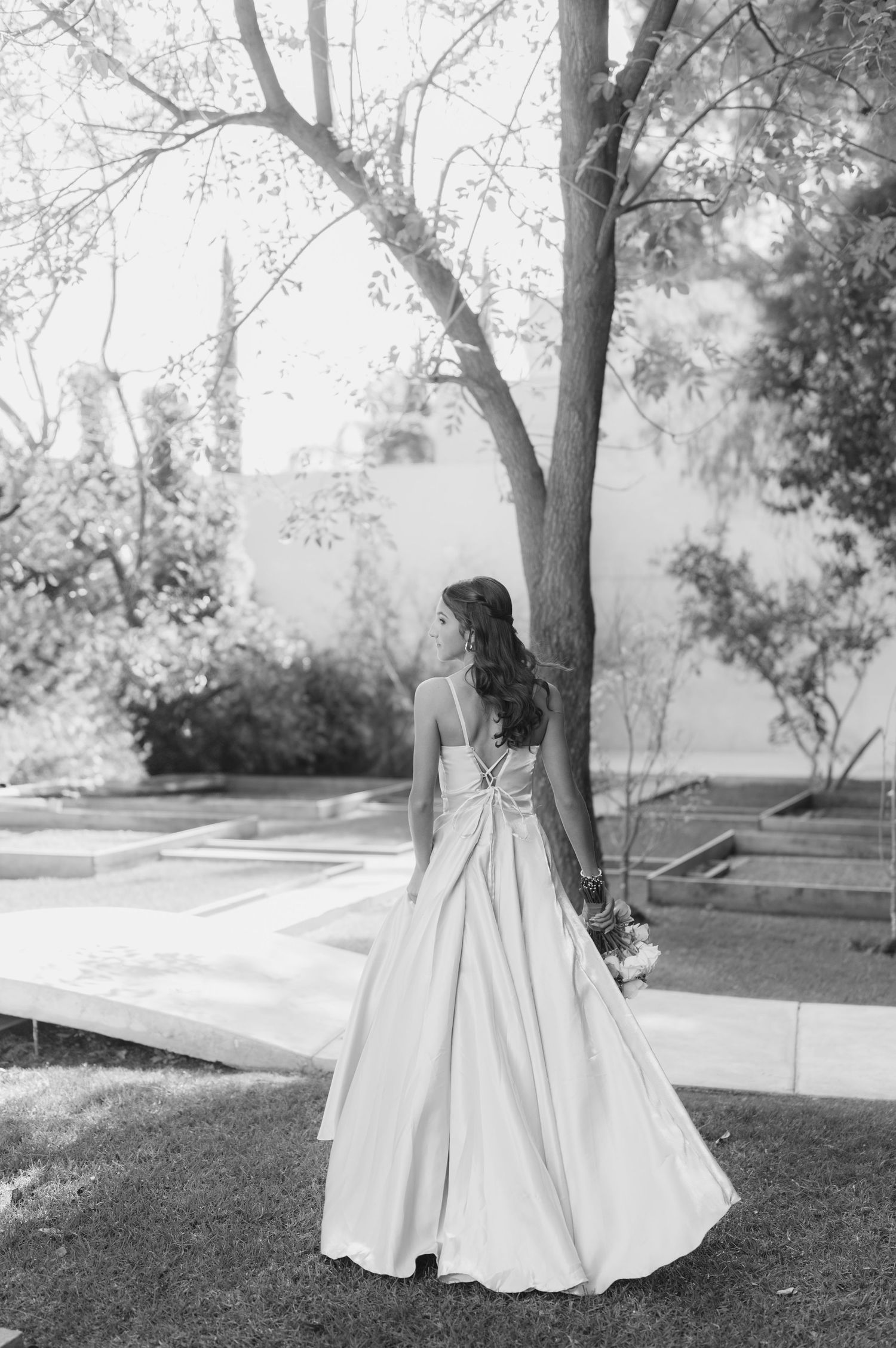 Black and white portrait of a bride in a classic gown standing beneath trees in a garden setting.