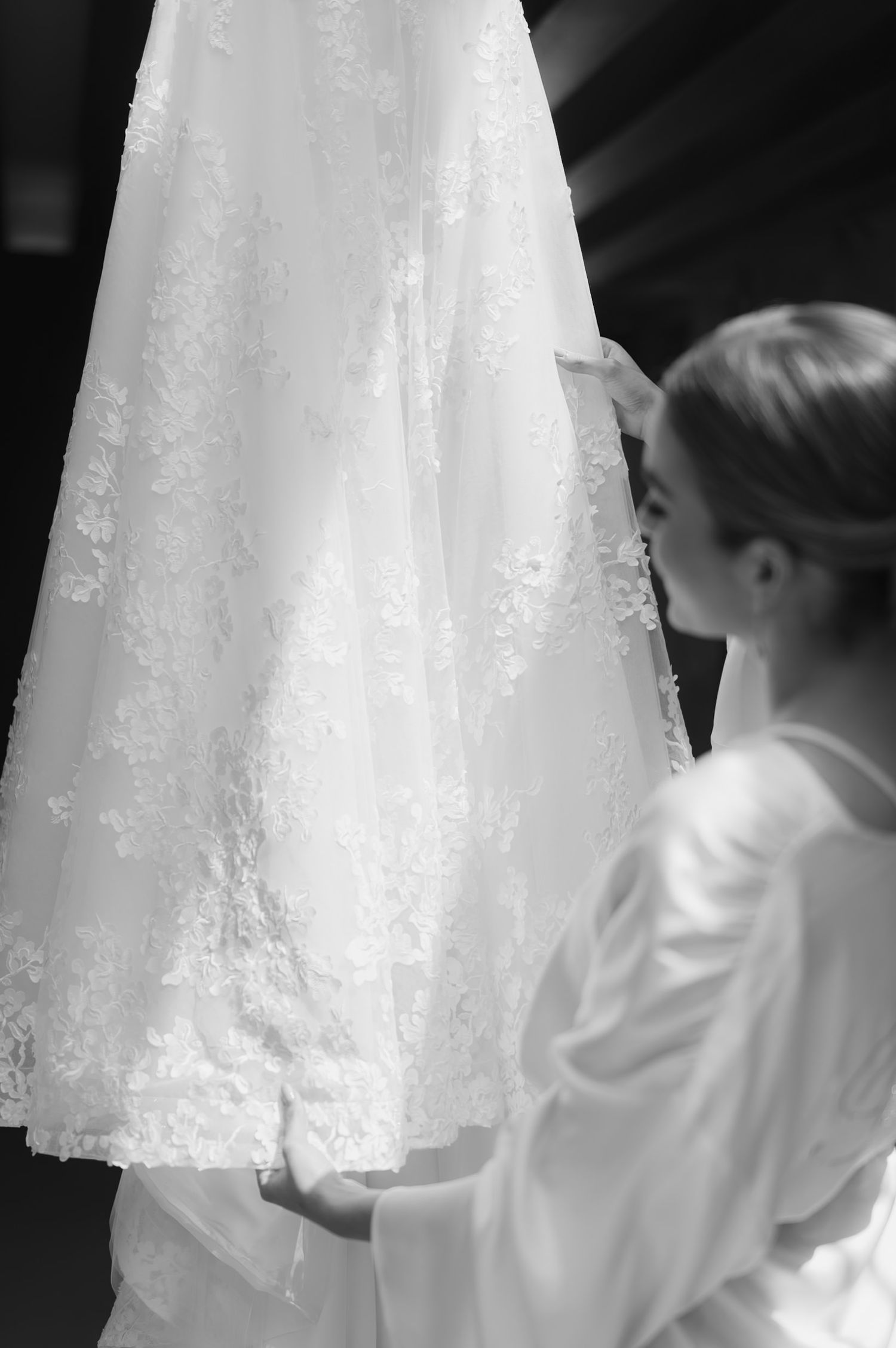 Black and white photo of a bride in a robe admiring her hanging lace wedding dress.