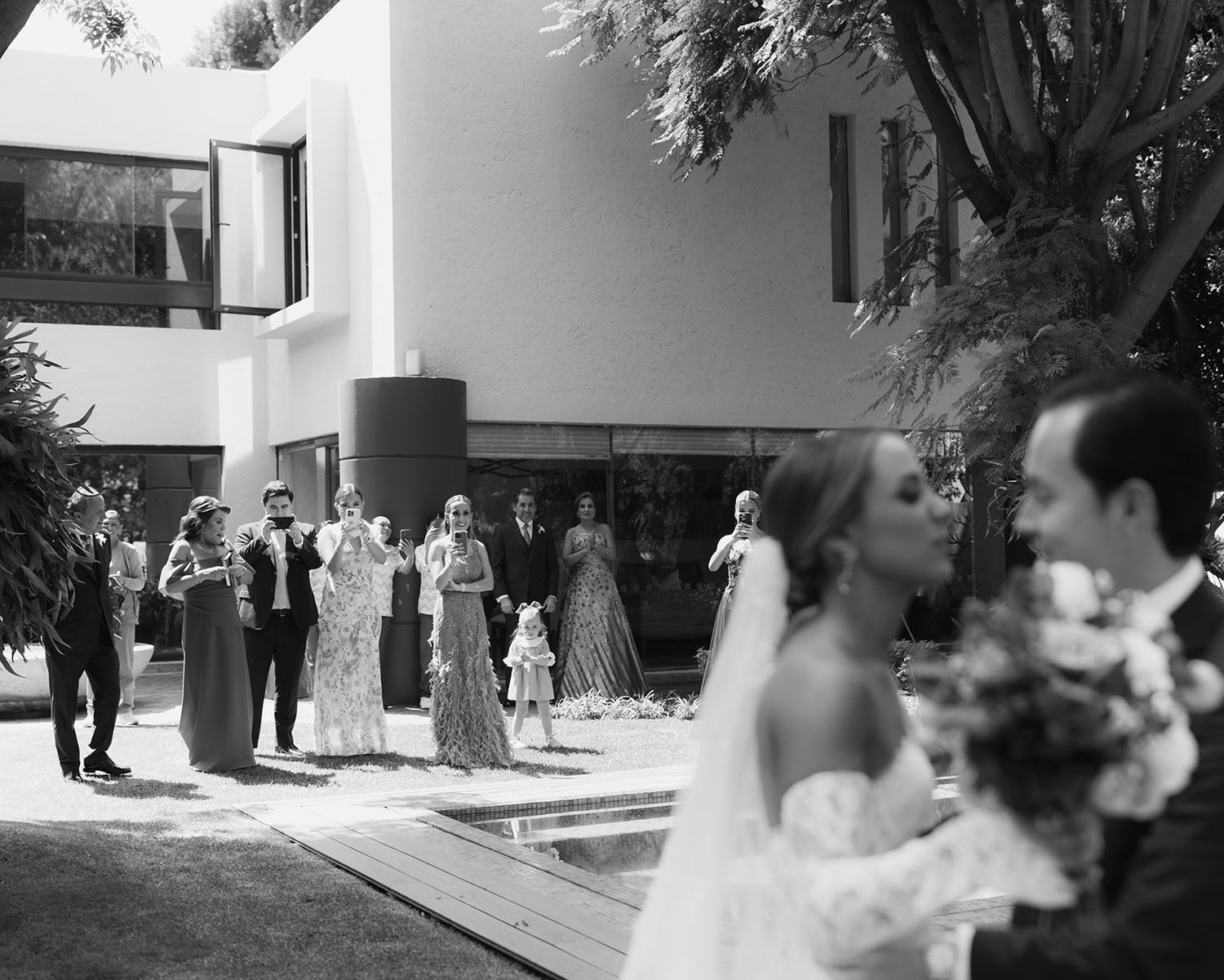 Black and white photo of wedding guests watching bride and groom embrace in a modern courtyard.