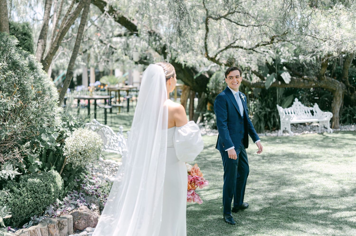Groom in navy suit turns during first look as bride in white gown and veil approaches.