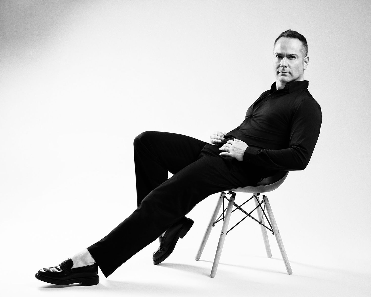 Man dressed in black sitting elegantly on a modern chair in a black and white studio shot.