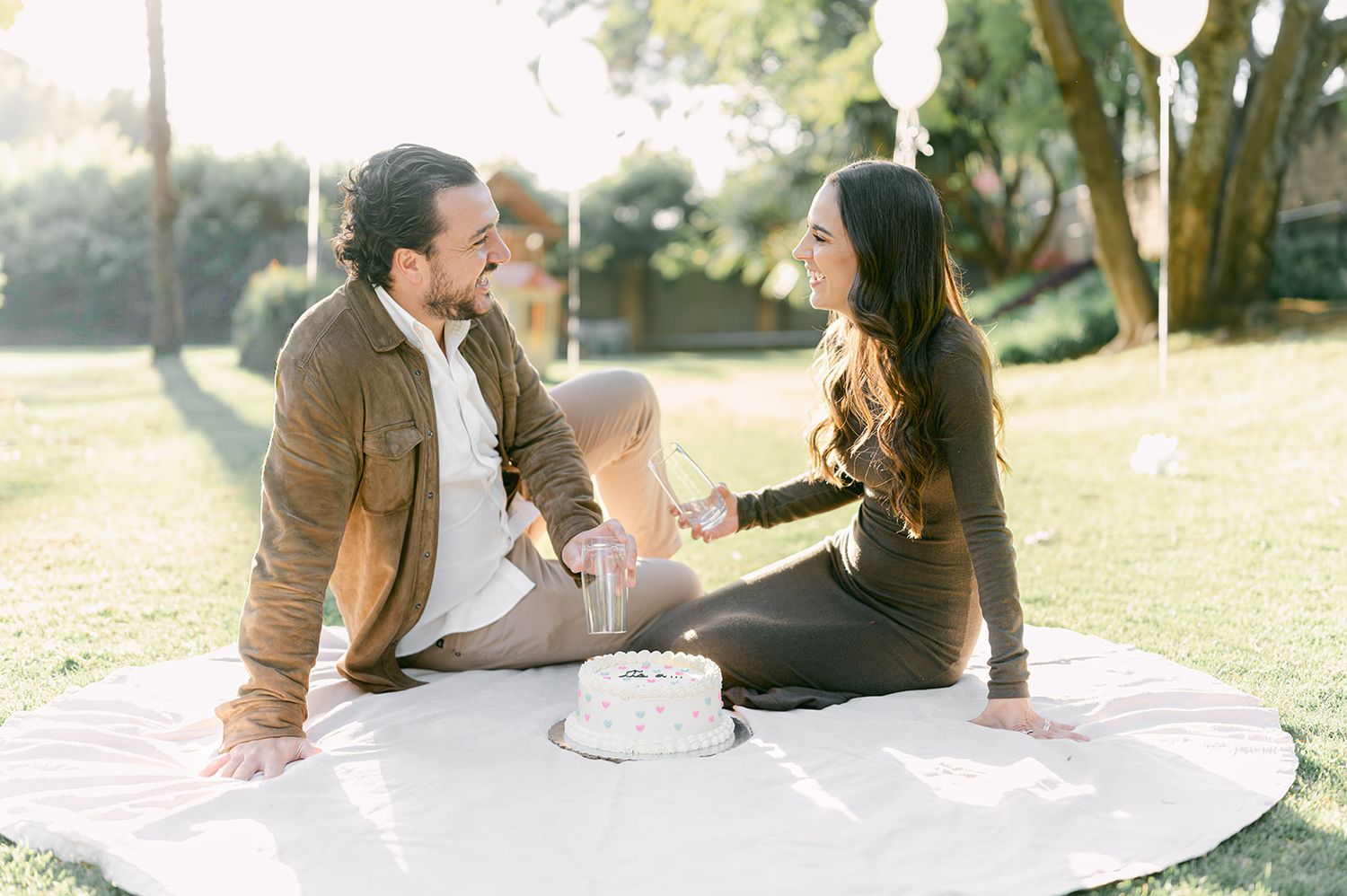 Smiling couple sitting together on white blanket in park with gender reveal cake in golden sunlight.