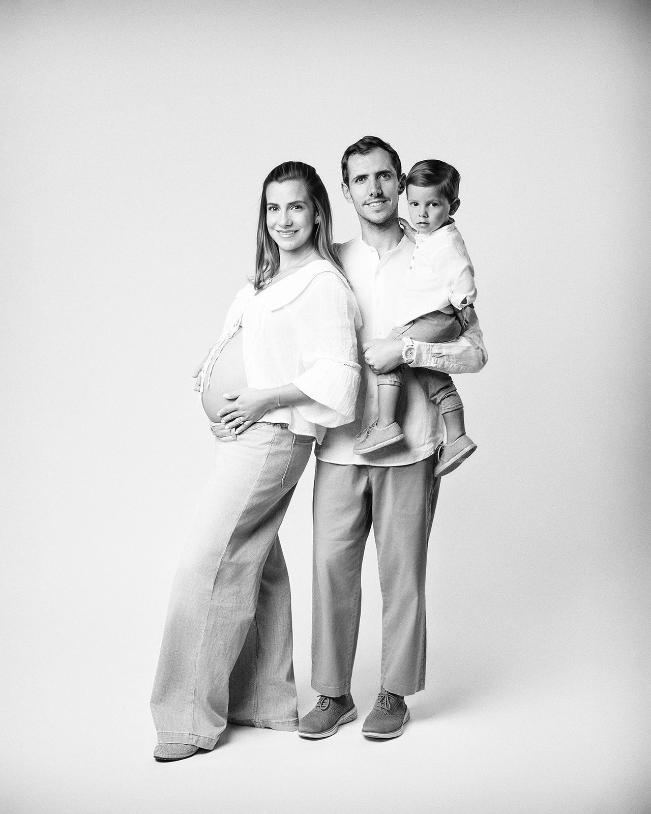 Black and white maternity photo of pregnant woman, man holding toddler, studio portrait of expecting family.