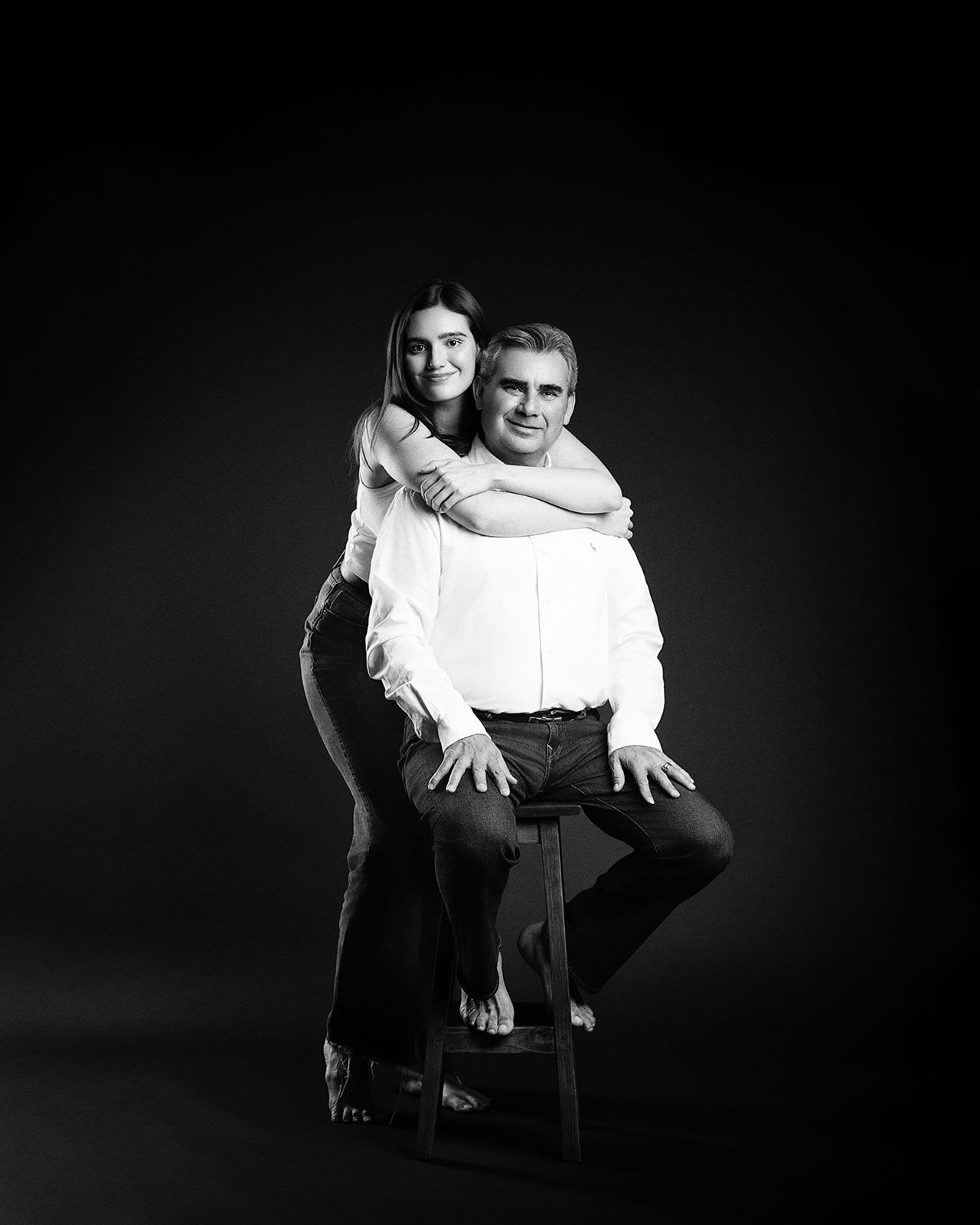 Black and white studio portrait of a smiling man seated on a stool with a young woman hugging him from behind.
