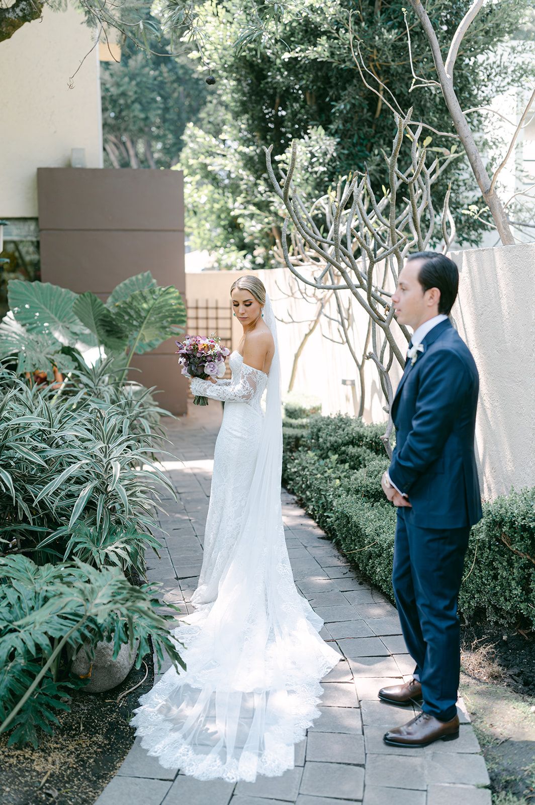 Bride in off-shoulder lace wedding gown with floral train stands with groom in navy suit during first look.