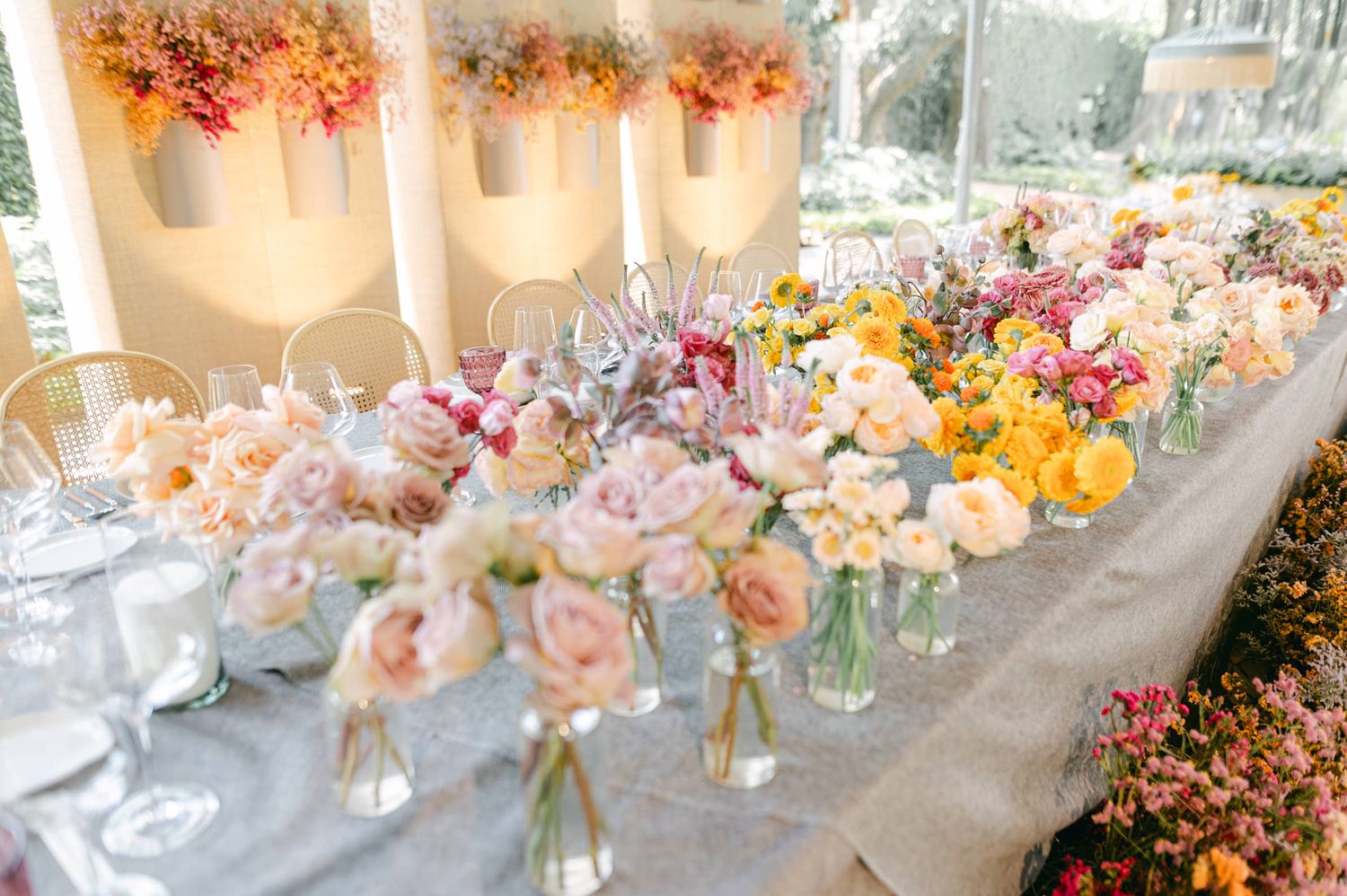 Long wedding reception table adorned with abundant pink, peach, and yellow floral arrangements in glass vases.