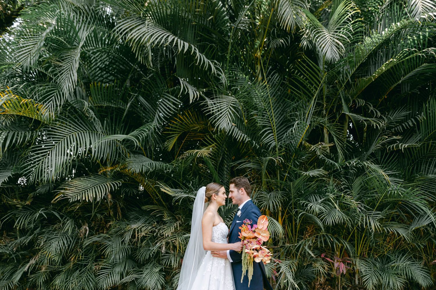 Bride and groom sharing a romantic moment surrounded by lush tropical palm foliage.