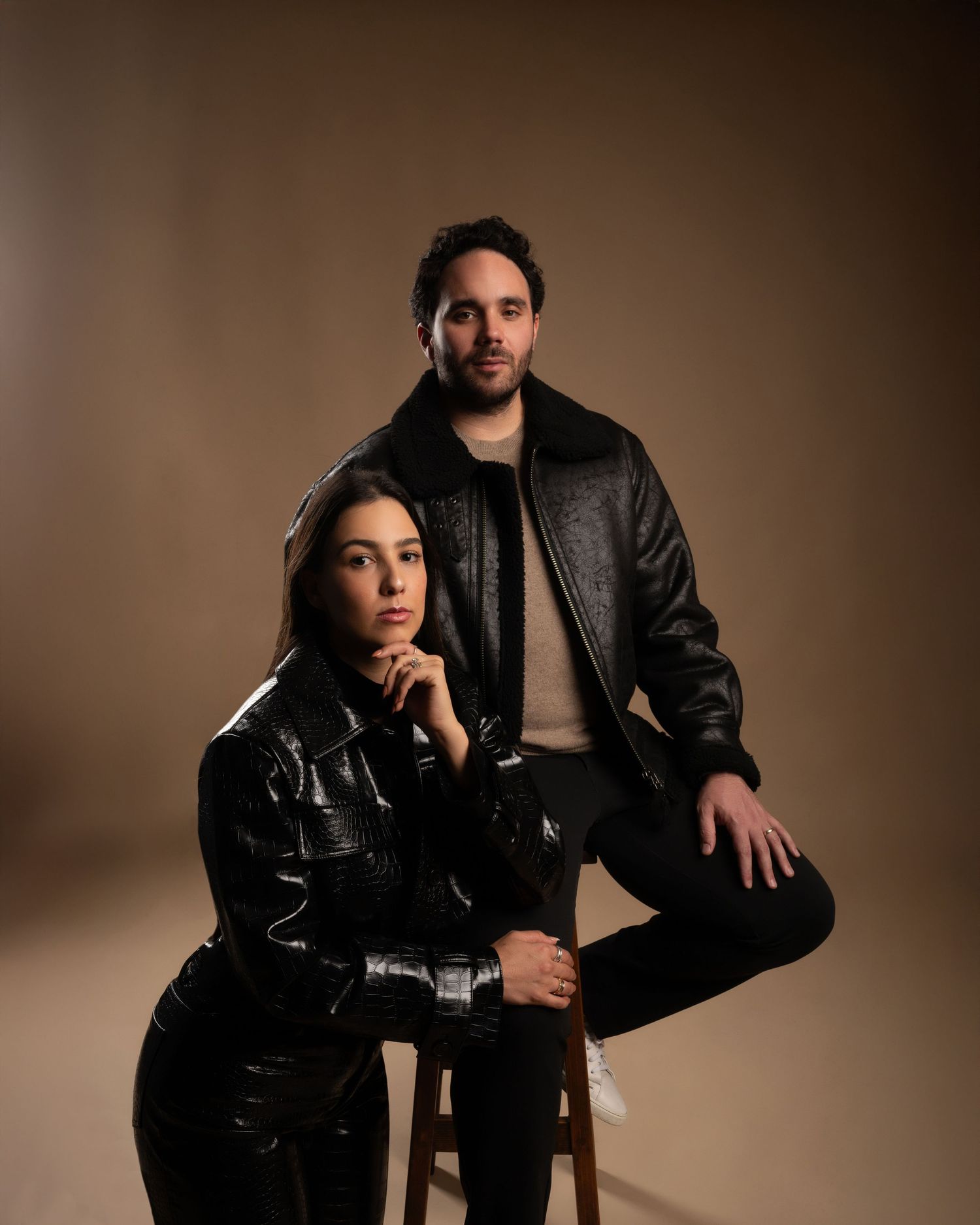 Woman in black patent leather jacket sits on stool while man in leather jacket stands behind in studio.