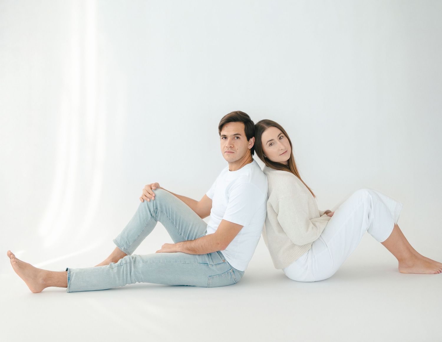 A couple sitting back-to-back on a white studio floor, dressed in casual white and cream outfits.