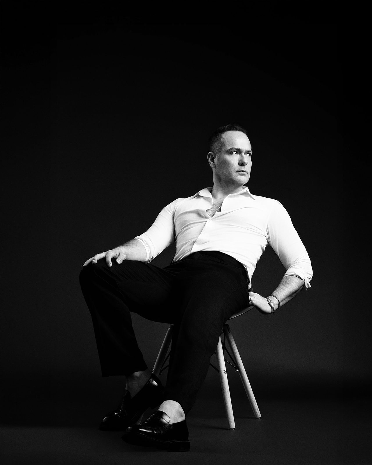 Black and white studio portrait of a man in white shirt and black trousers seated on a stool, looking away confidently.