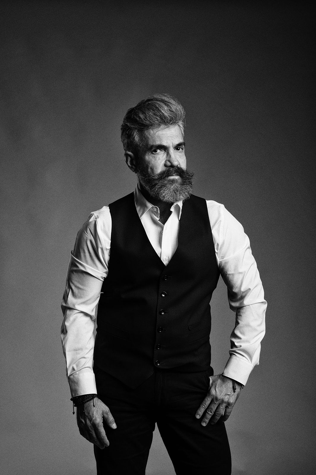 Black and white portrait of a bearded man in a white shirt and dark vest posing confidently against a gray background.