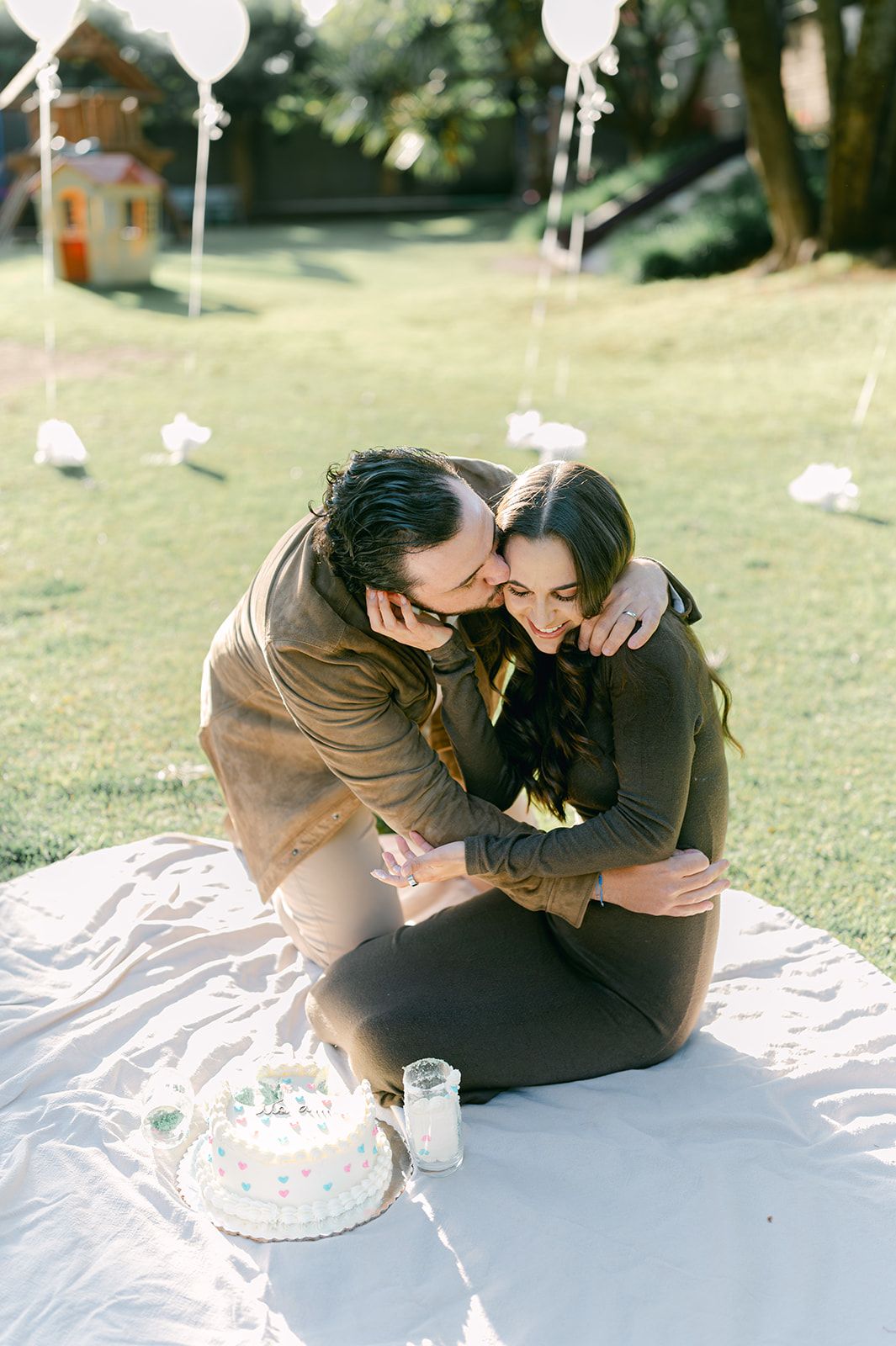 Couple sitting on blanket in park celebrating with cake and balloons at gender reveal party.