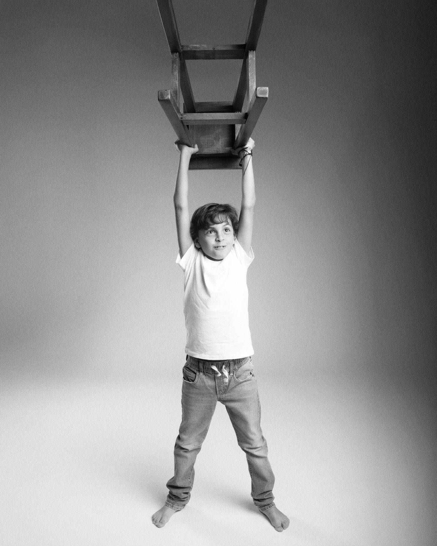 Black and white photo of a young boy lifting a wooden chair above his head in a studio.