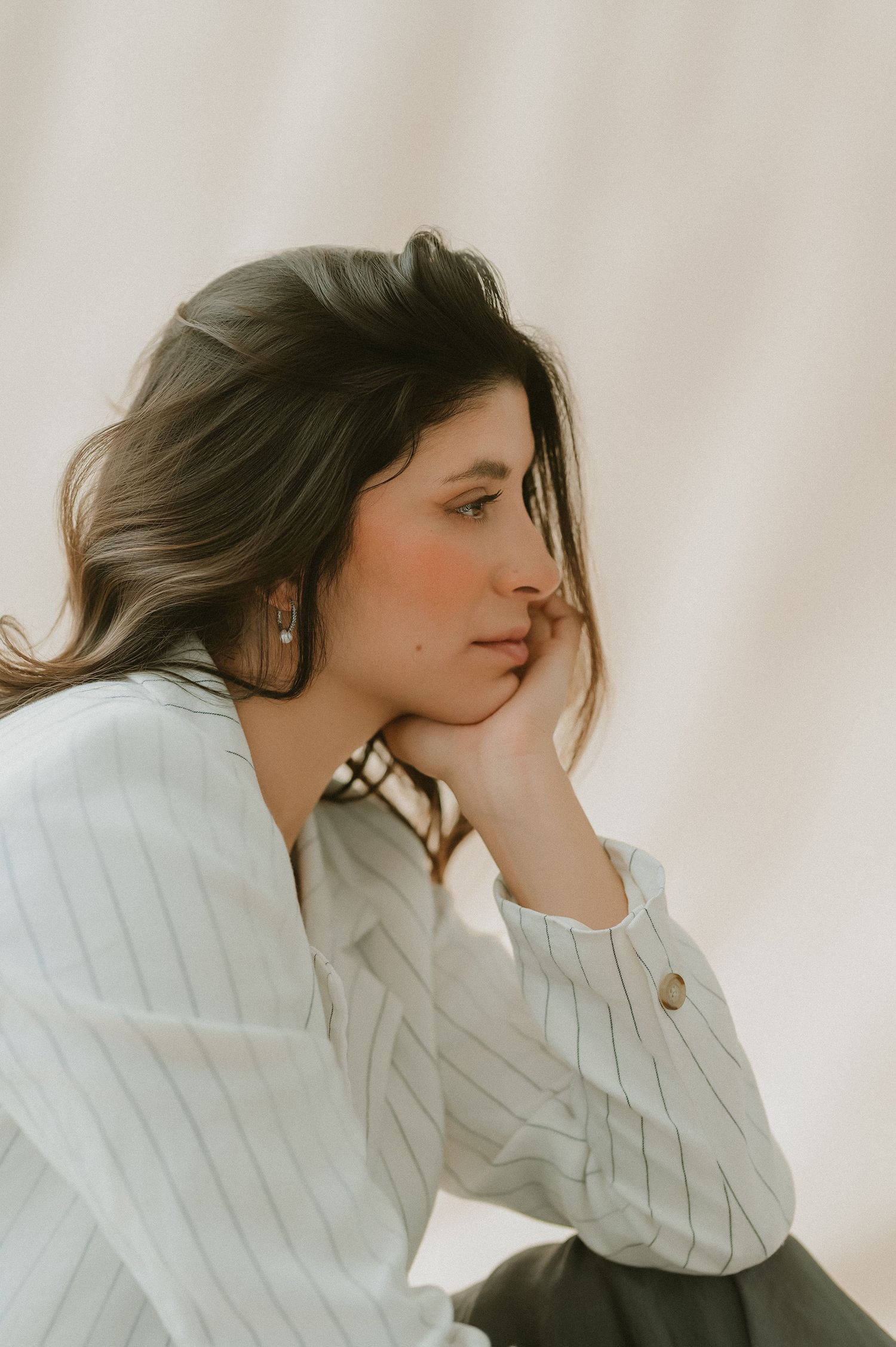 Close-up of brunette woman in striped white blazer resting chin on hand, contemplative side profile portrait.
