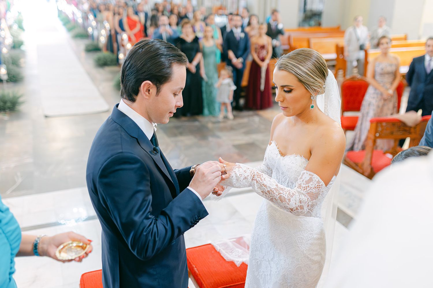 Groom in navy suit places wedding ring on bride's finger during church ceremony.