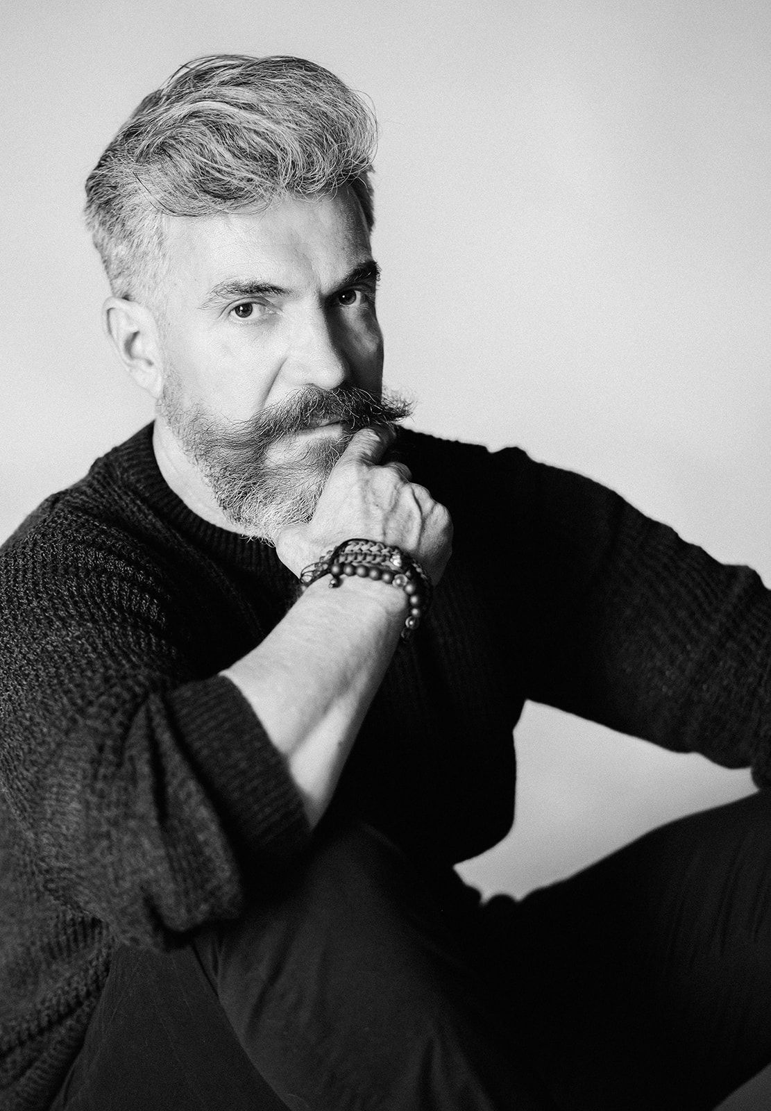 Black and white portrait of a bearded man with gray hair wearing a dark sweater and beaded bracelet.