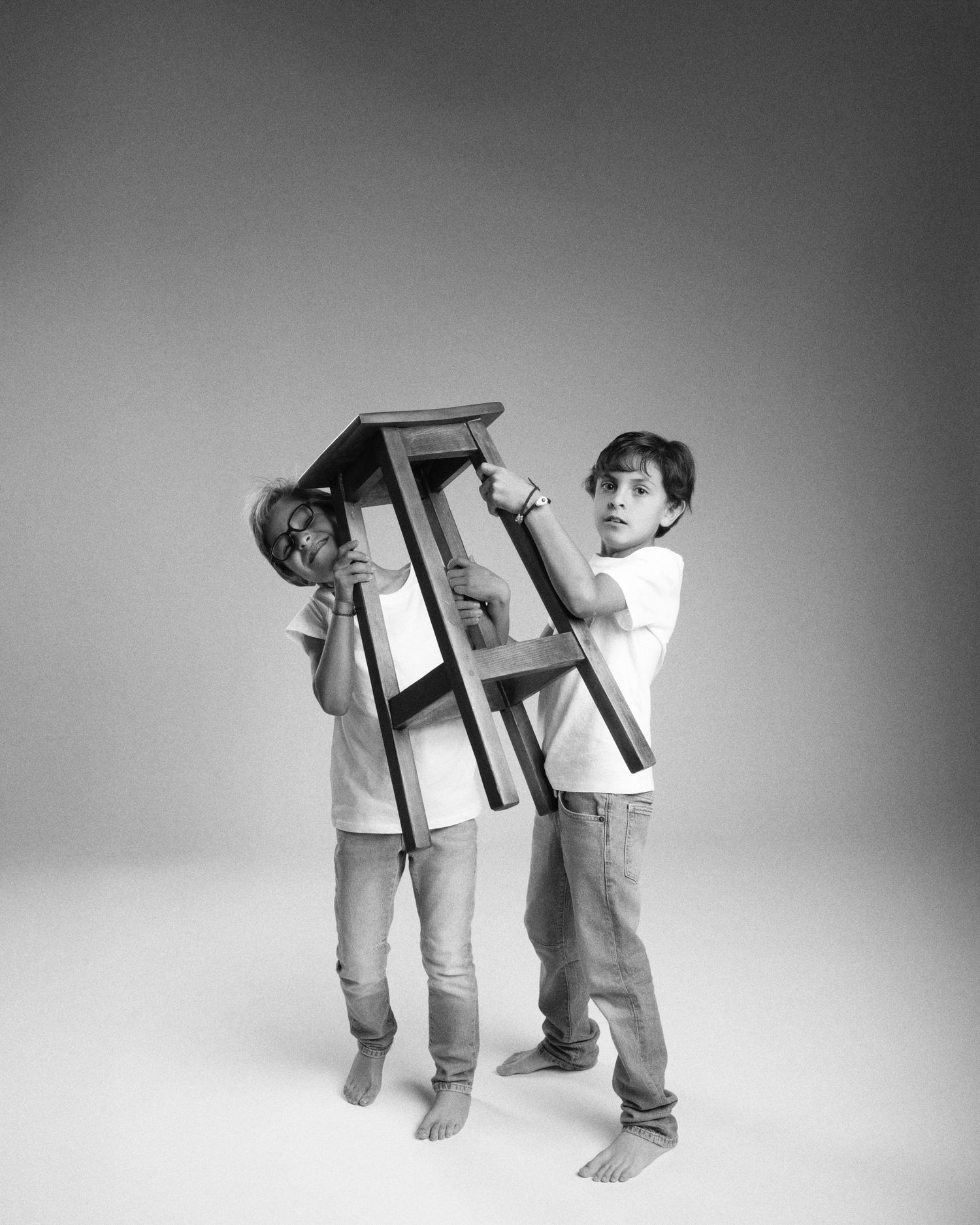 Black and white photo of two boys playfully lifting a wooden stool in a photography studio.