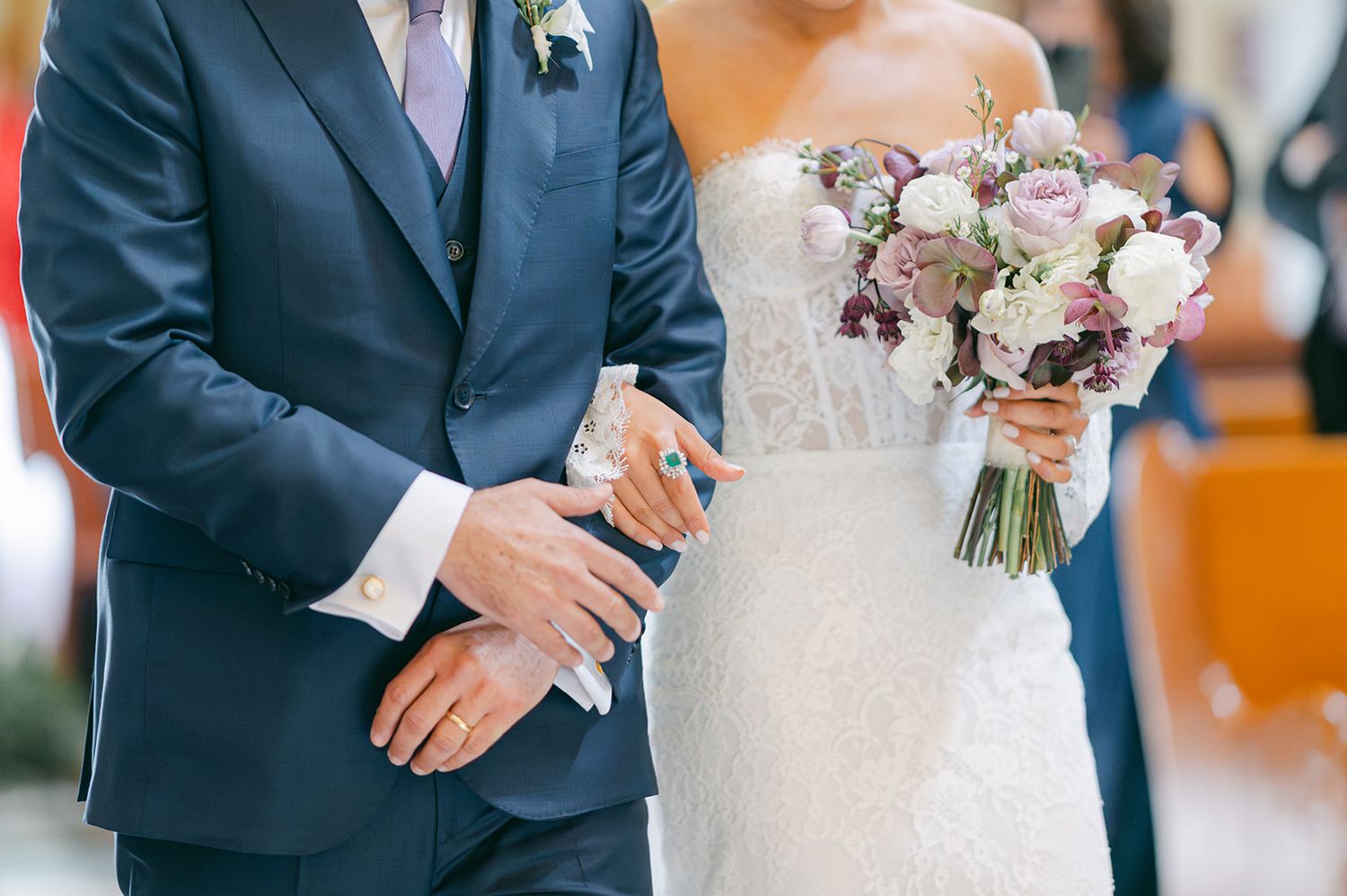 Close-up of bride and groom walking arm in arm at ceremony, bride holding purple and white floral bouquet.