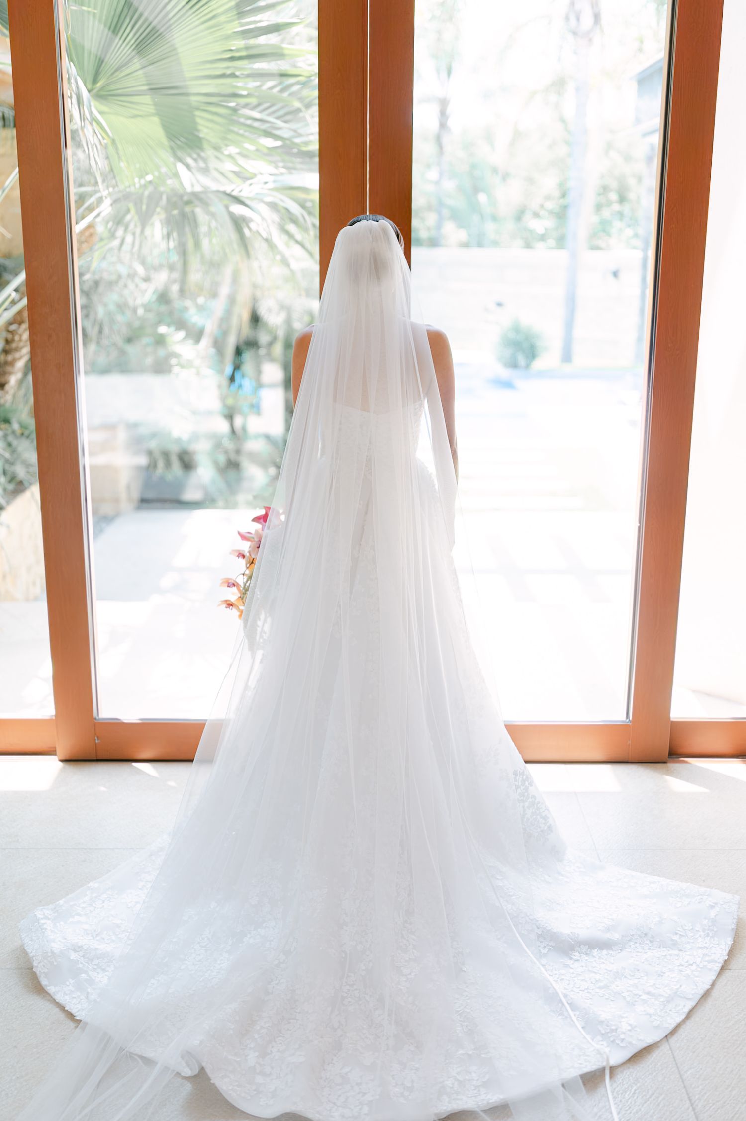 Bride in a stunning lace wedding gown with cathedral veil standing before large windows overlooking lush greenery.