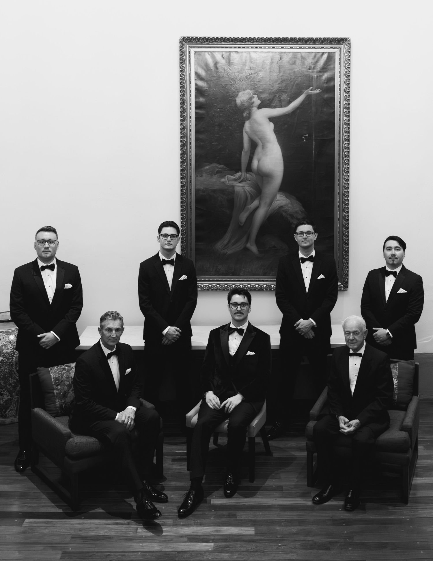 Black and white wedding party portrait of groomsmen in tuxedos posed before a classical painting.