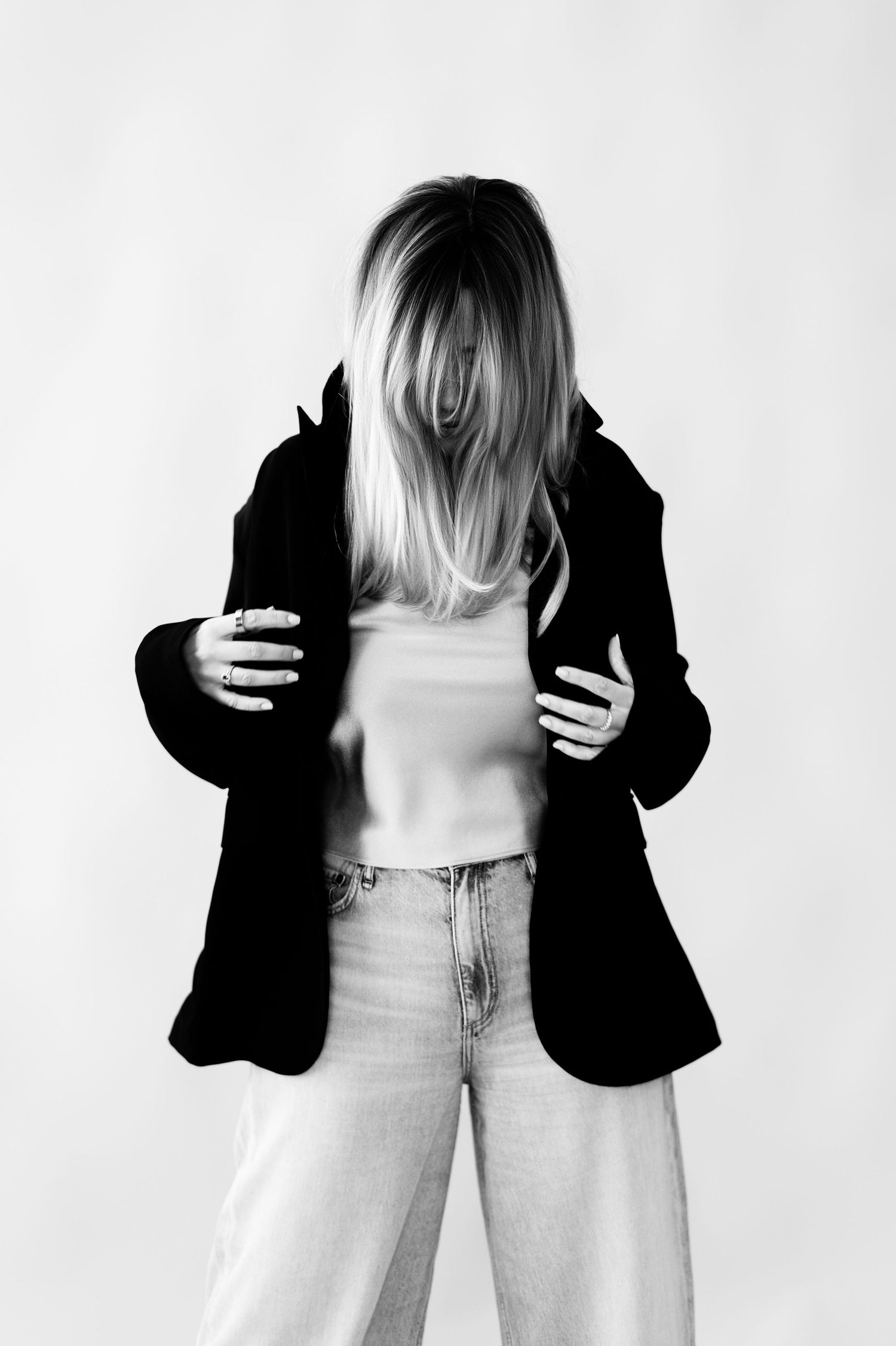 Black and white studio photo of a blonde woman in blazer and jeans with hair covering face.