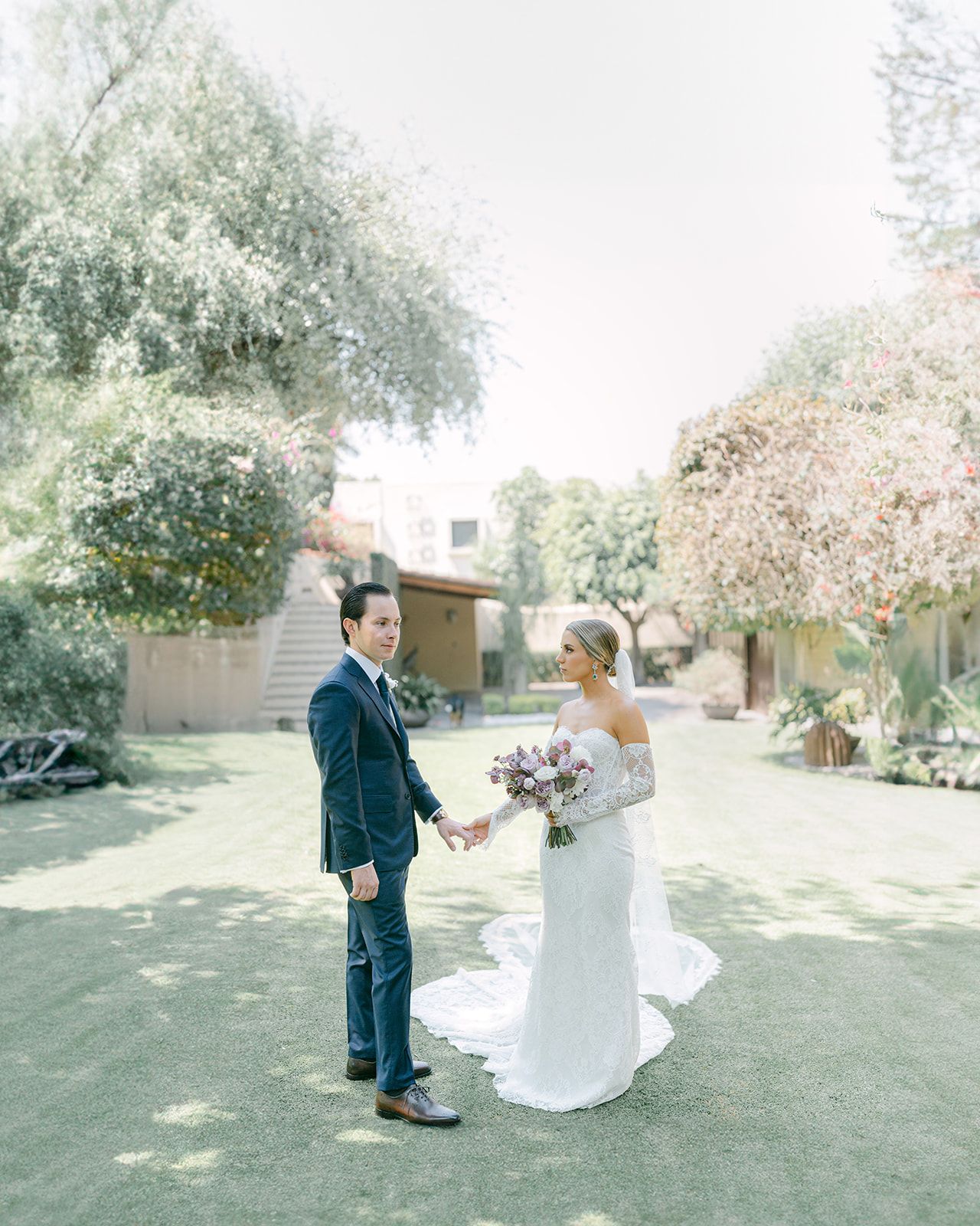 Bride and groom holding hands in lush garden, bride wearing lace off-shoulder wedding gown with veil.